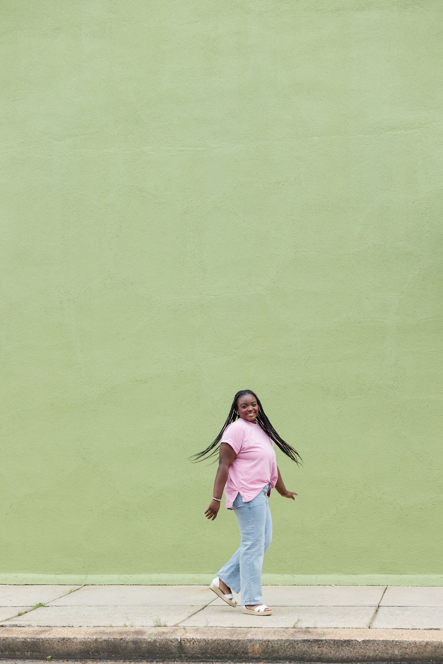 A joyful young Black woman with long braids walks along a sidewalk in a pink top and blue jeans against a green wall.