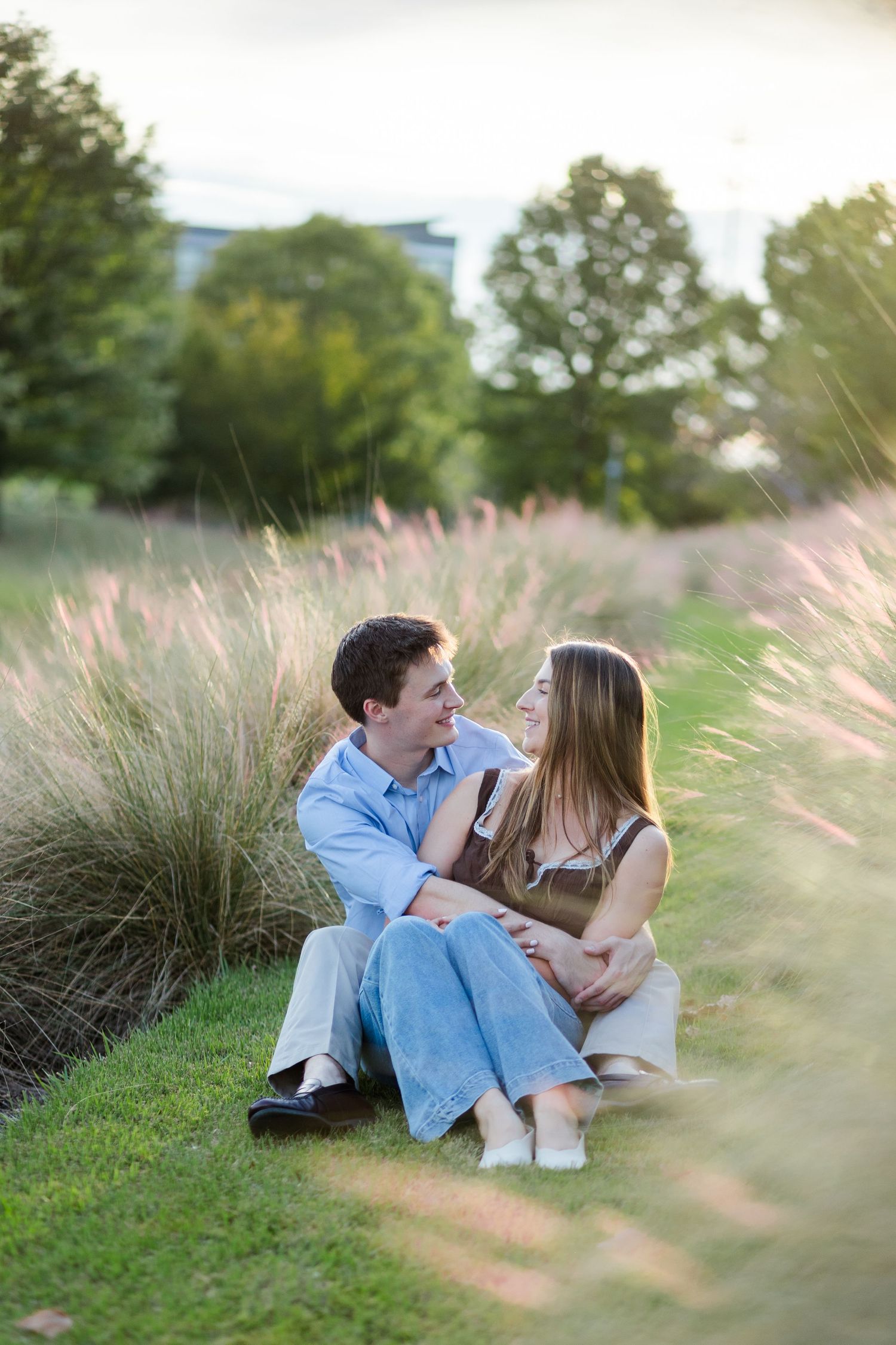 Happy couple sitting together on green grass beside tall ornamental grasses during golden hour outdoors.