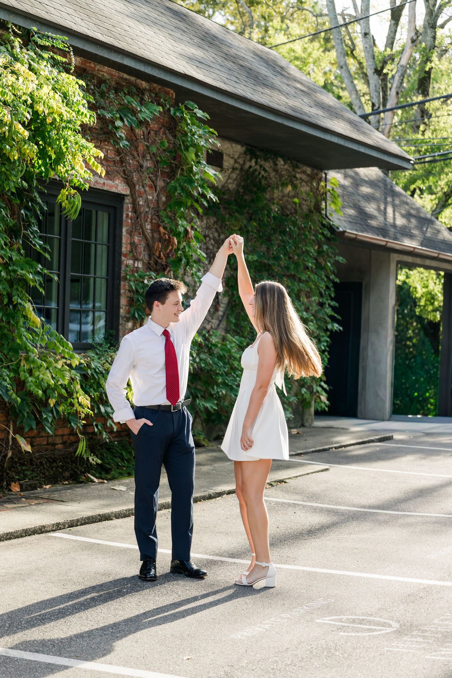 Couple dancing outdoors, man in white shirt and red tie spinning woman in white dress in sunny parking lot.