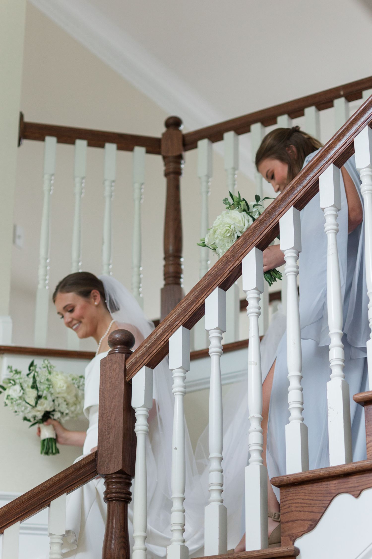 Bride and bridesmaid in light blue dresses descend elegant wooden staircase holding white floral bouquets.