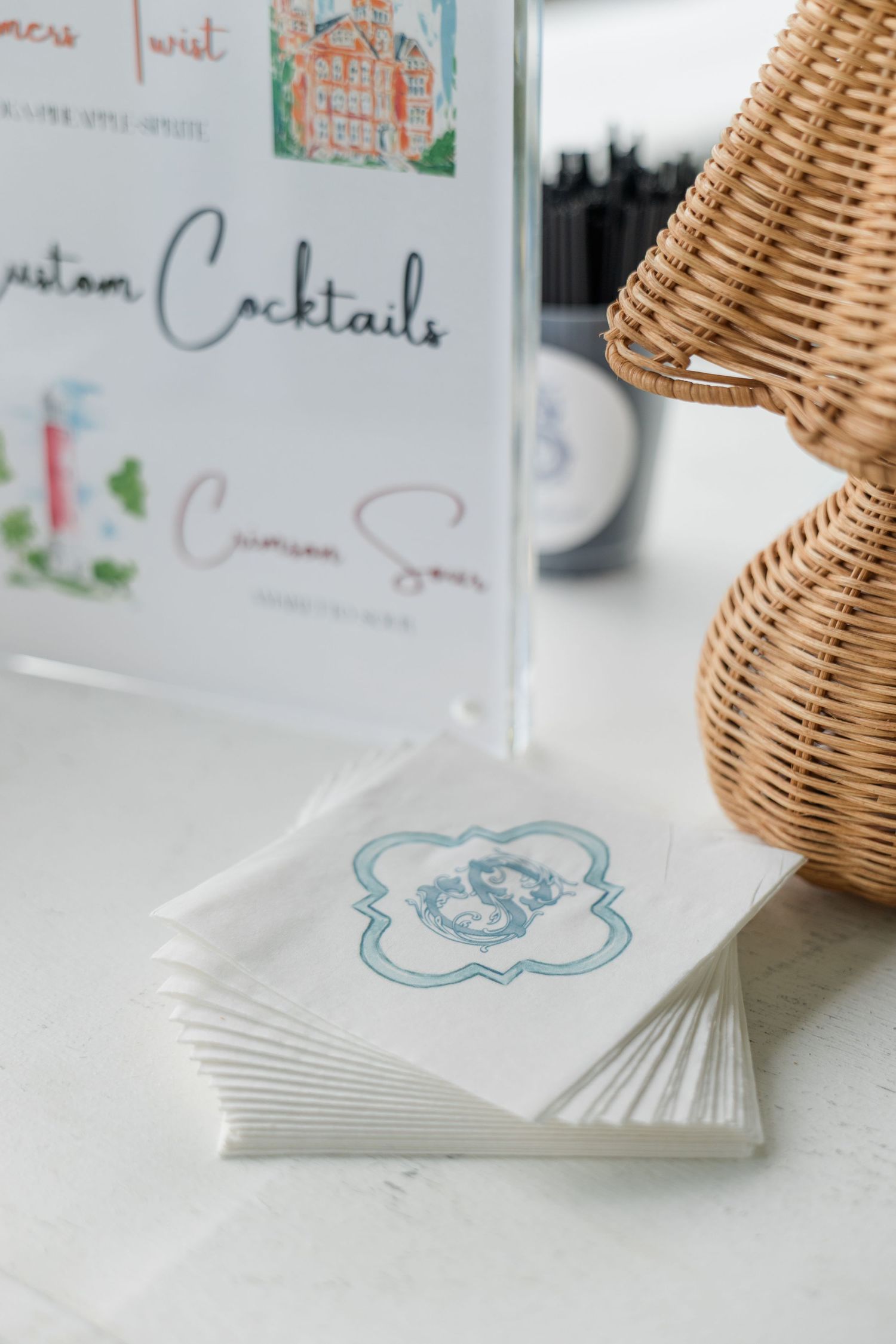 Monogrammed cocktail napkins stacked beside a custom cocktails sign and wicker basket at a wedding bar.