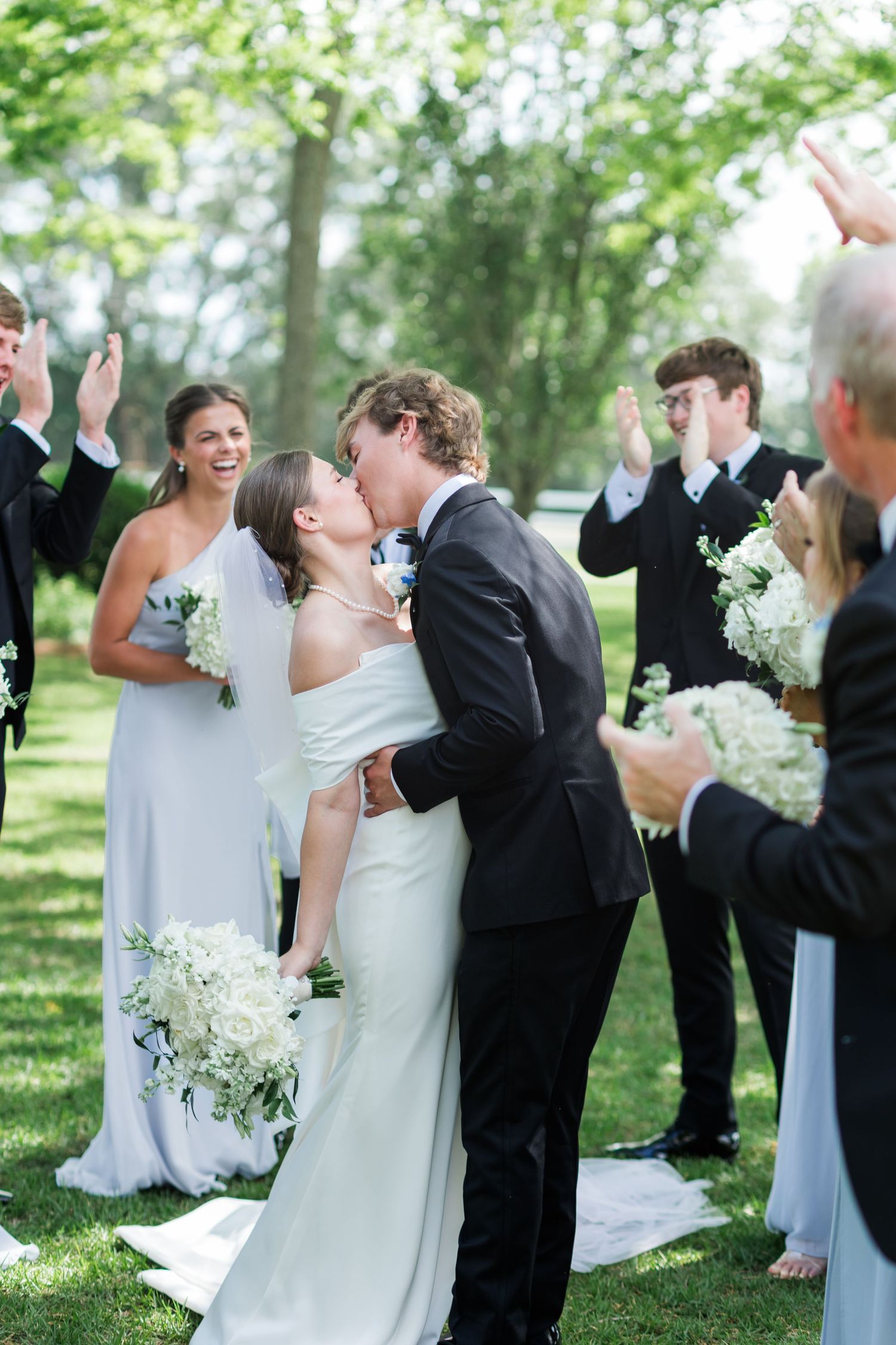 Bride and groom share first kiss at outdoor wedding ceremony surrounded by cheering guests and wedding party.