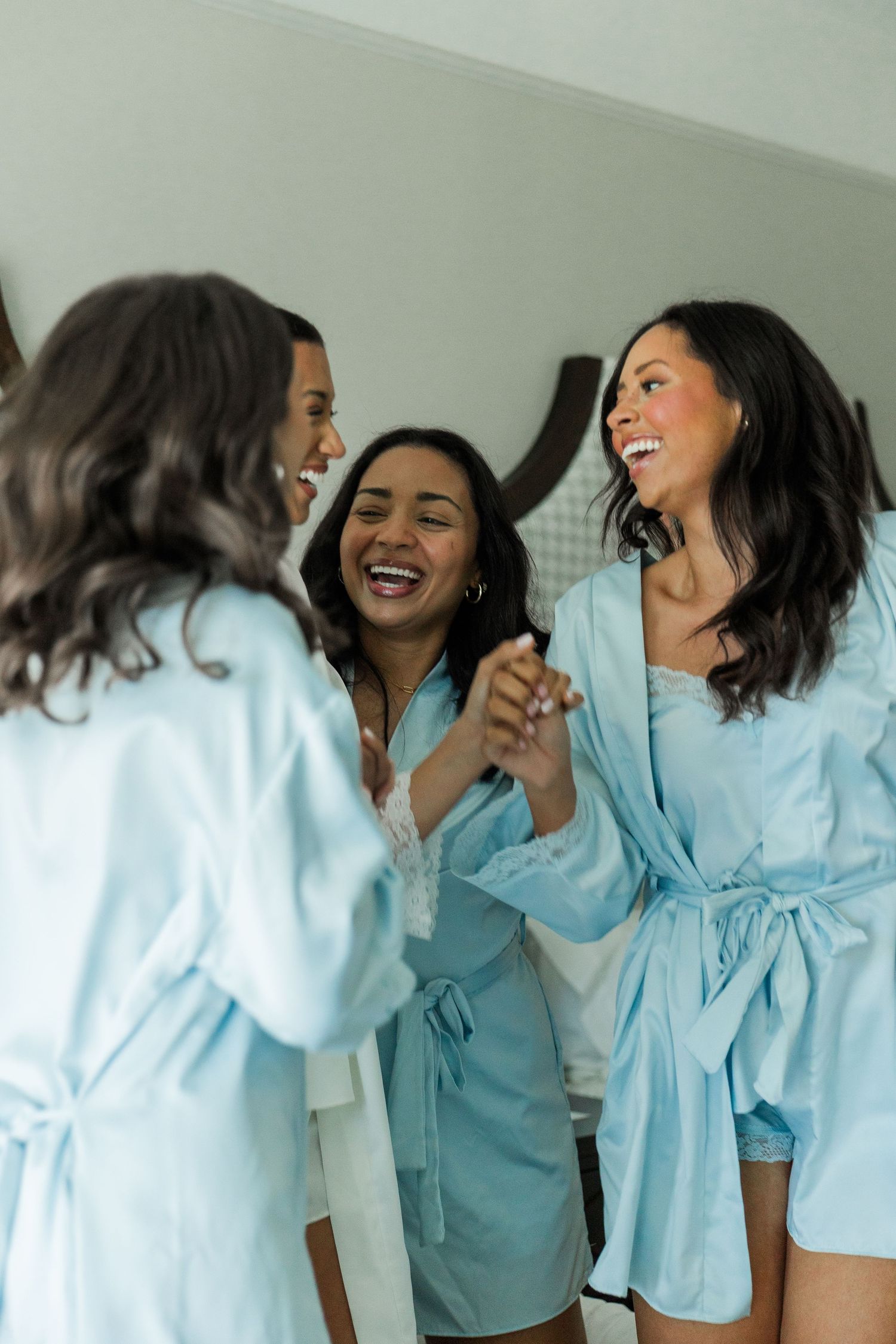 Three bridesmaids laughing and holding hands in matching light blue satin robes during wedding morning preparations.