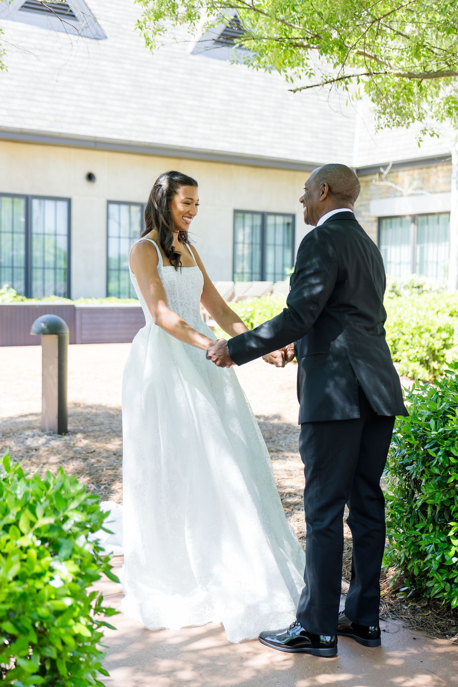 Bride in white gown and groom in black suit hold hands outdoors, smiling at each other on their wedding day.