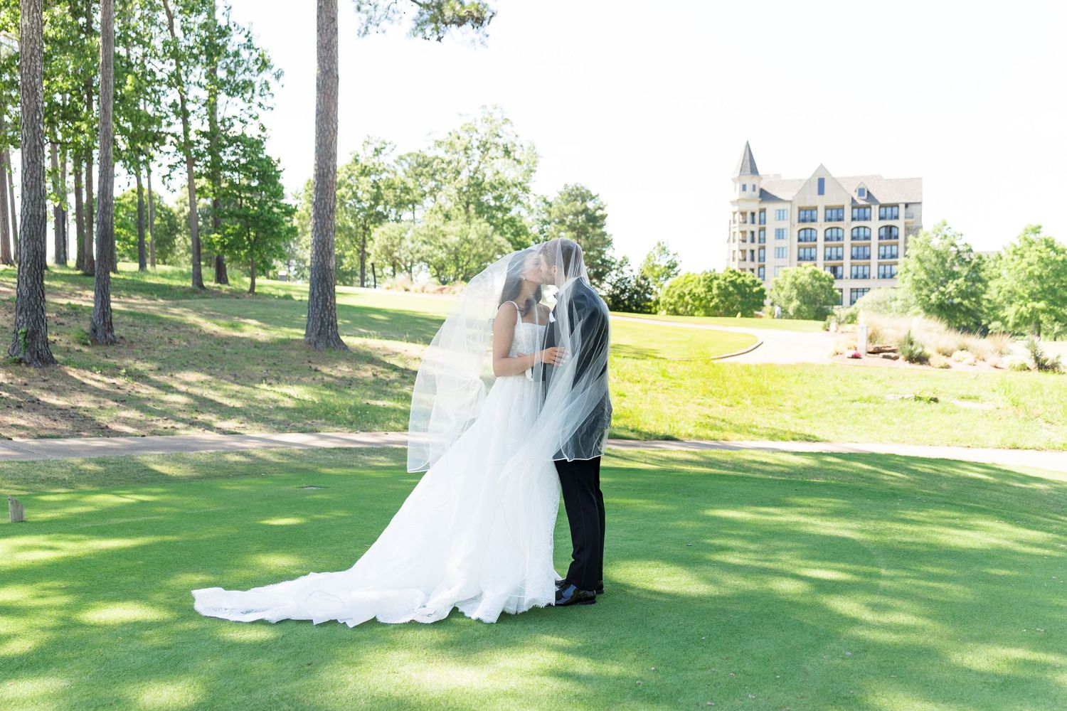 Bride and groom sharing a kiss under a flowing veil on a lush green golf course with a elegant venue in the background.