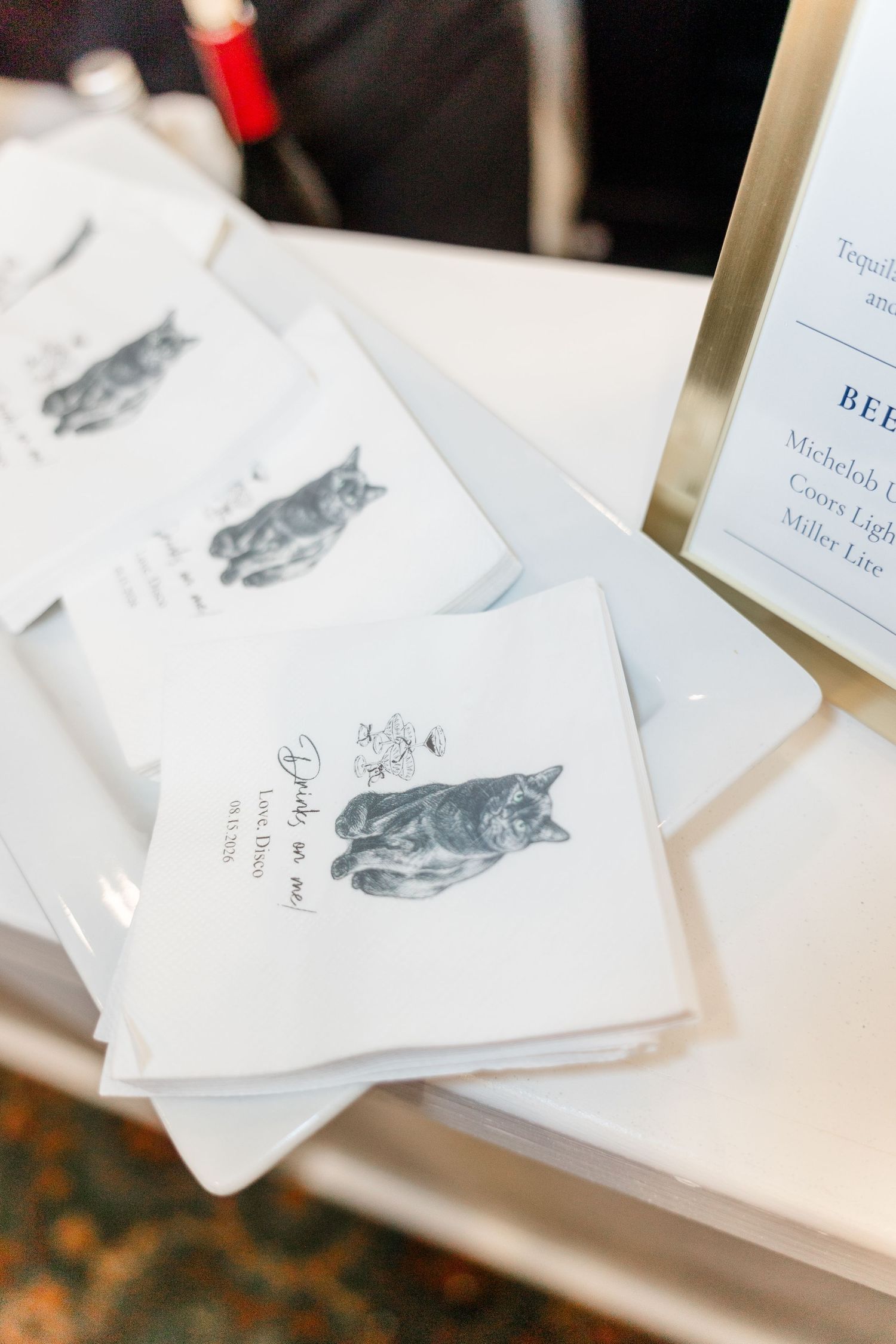 Custom wedding cocktail napkins featuring cat illustrations and couple's names displayed on a white table at a reception.