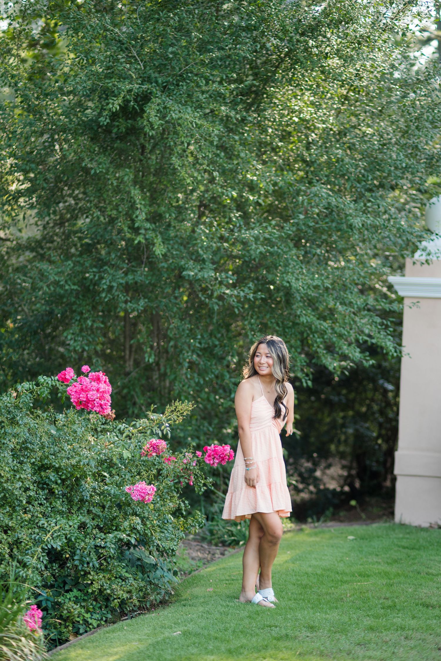 Smiling young woman in pink dress posing near vibrant pink blooming bushes in garden.