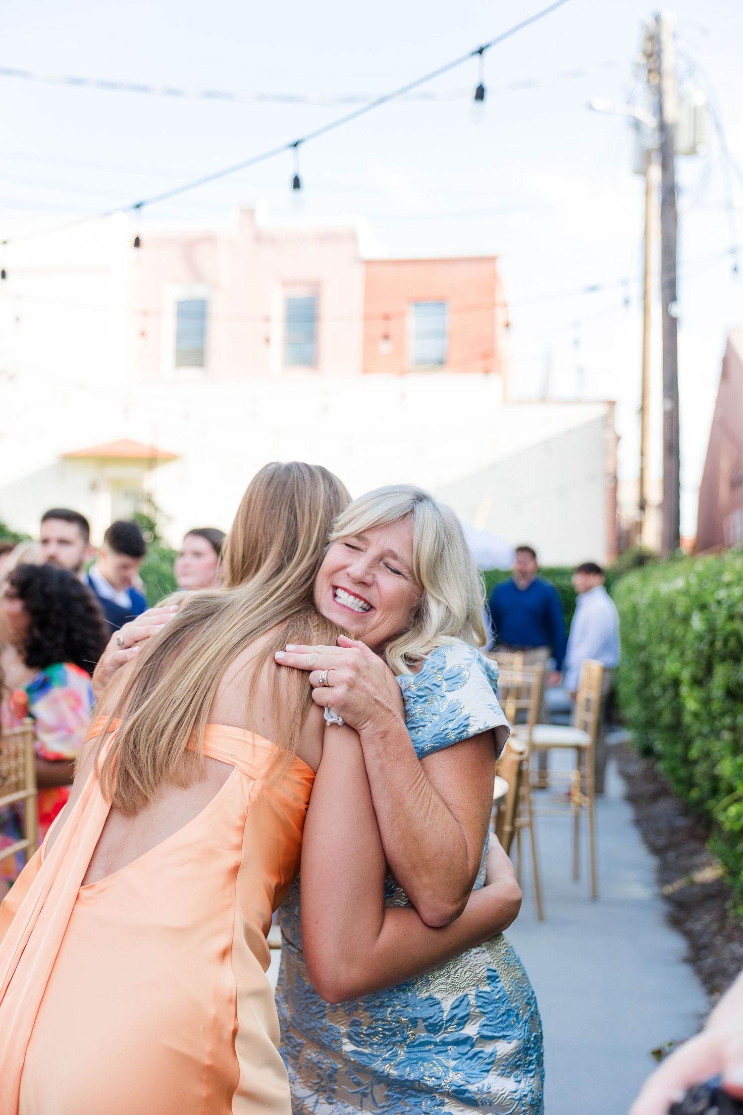 Two women share a warm embrace at an outdoor wedding reception under string lights.