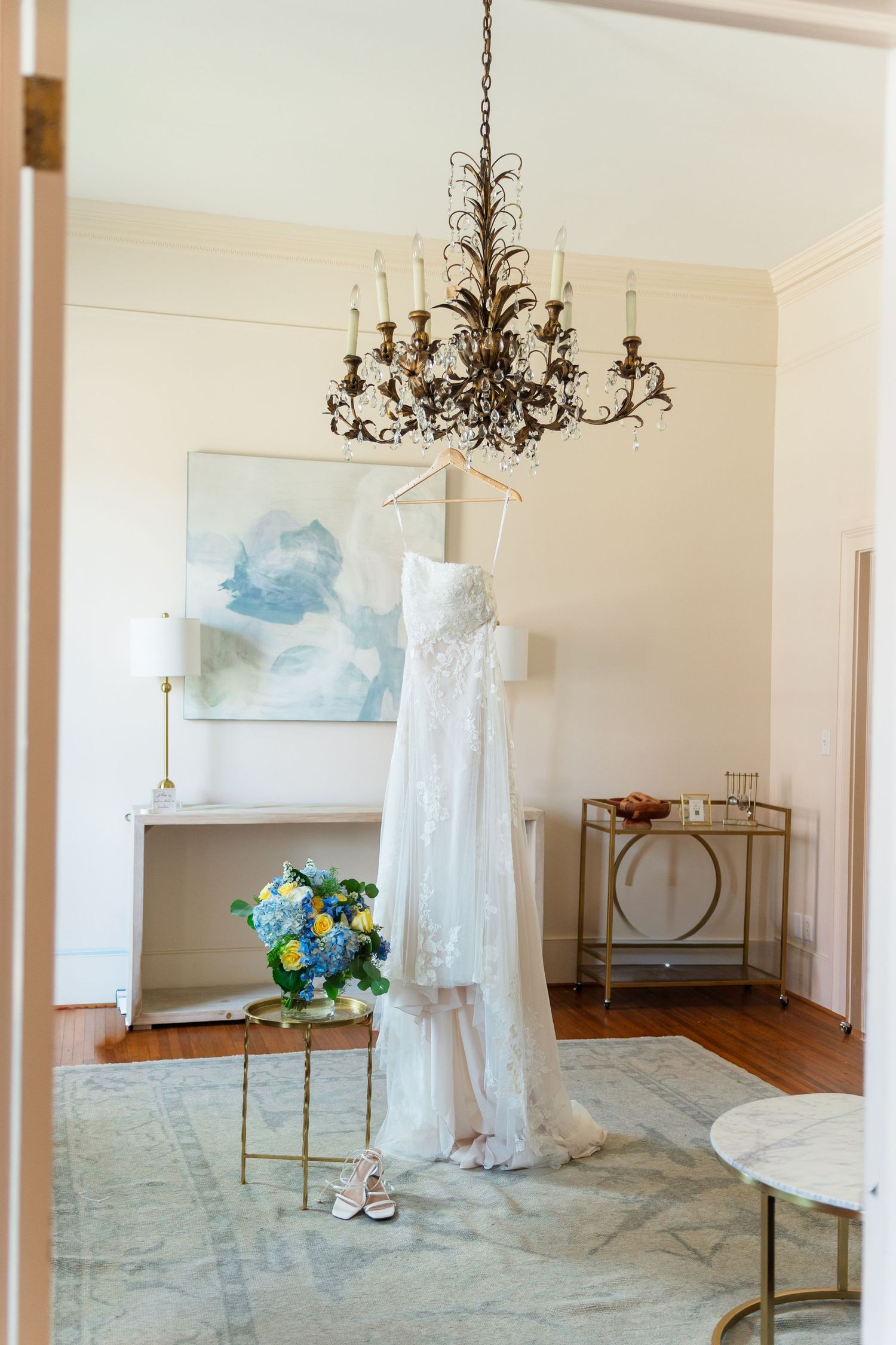 White wedding dress hanging from chandelier with blue floral bouquet in elegant bridal suite.