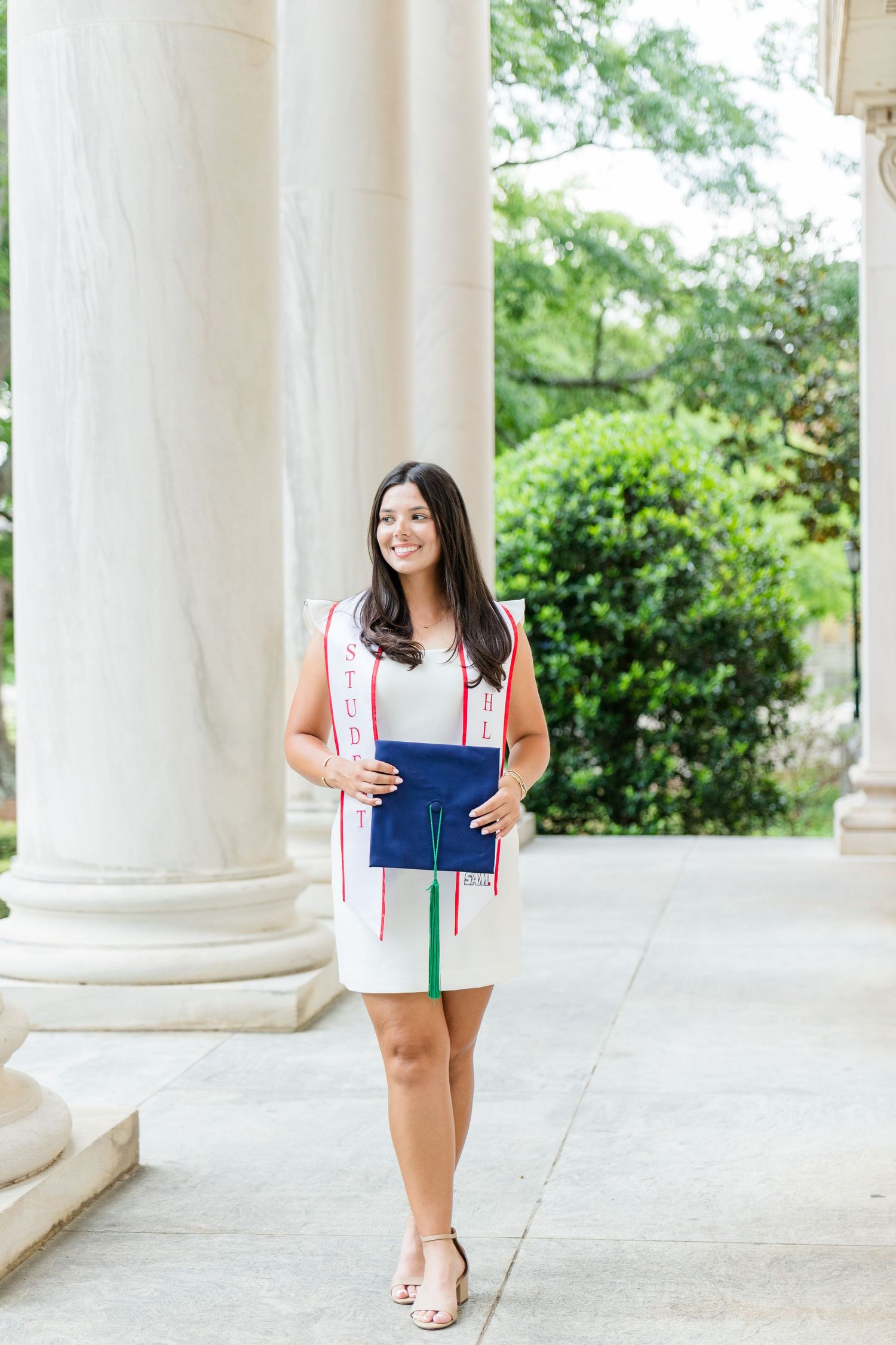 Female graduate standing between columns wearing a white dress and colorful stole, holding blue cap.