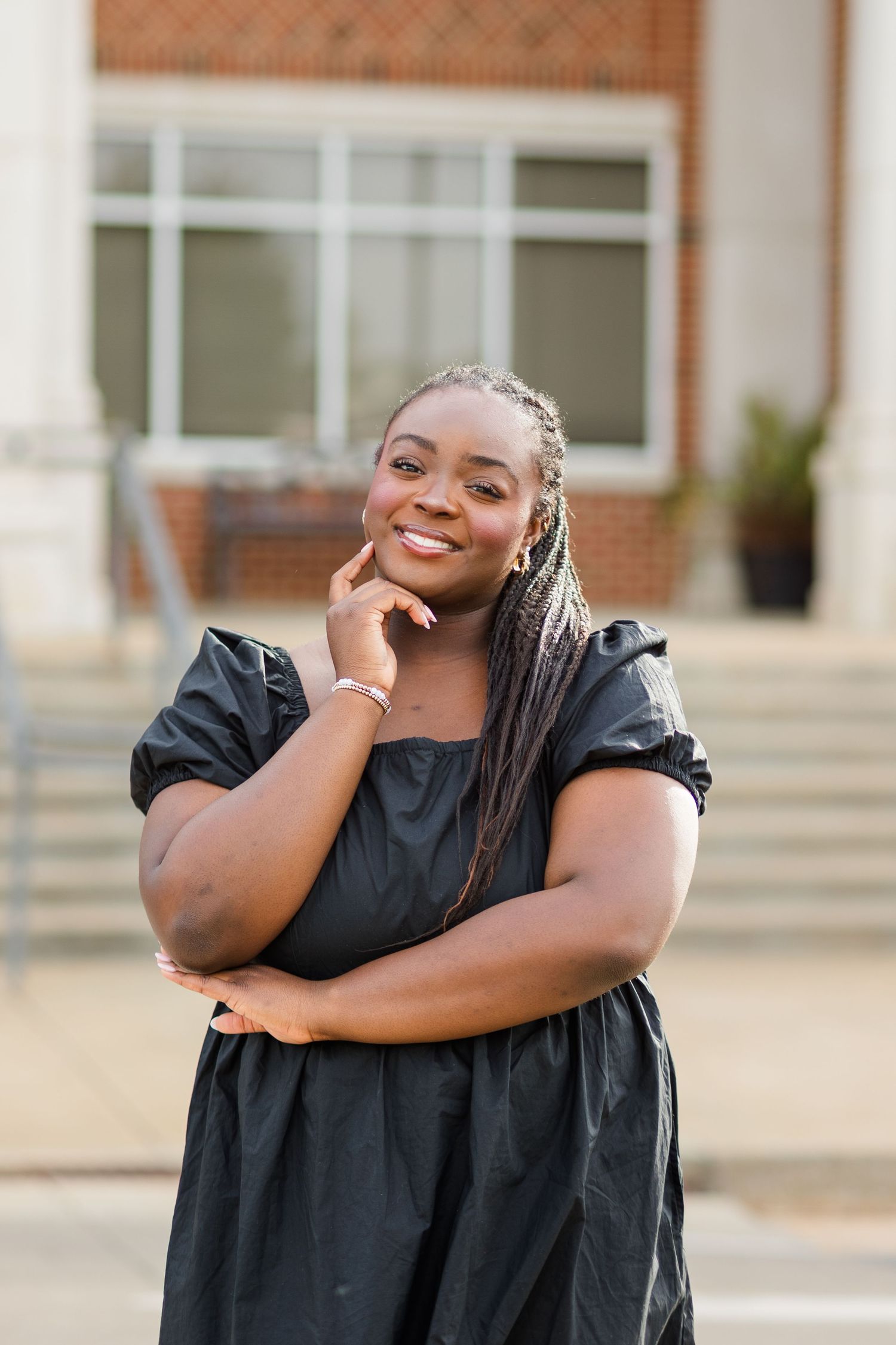 Young Black woman with braids smiling confidently in black dress, posing outdoors near brick building.