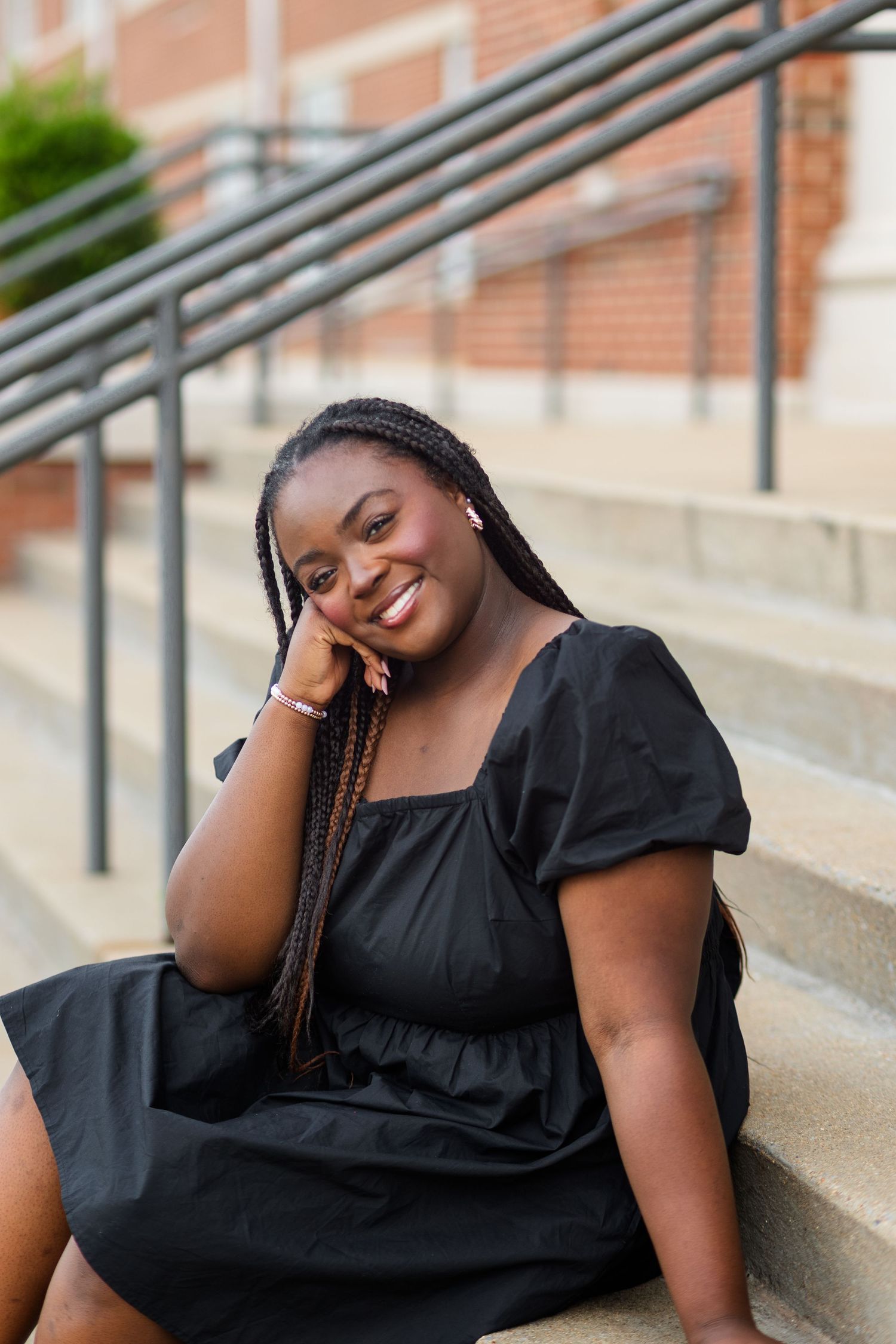 A young Black woman with braids sits on steps in a black dress, smiling warmly at the camera outdoors.