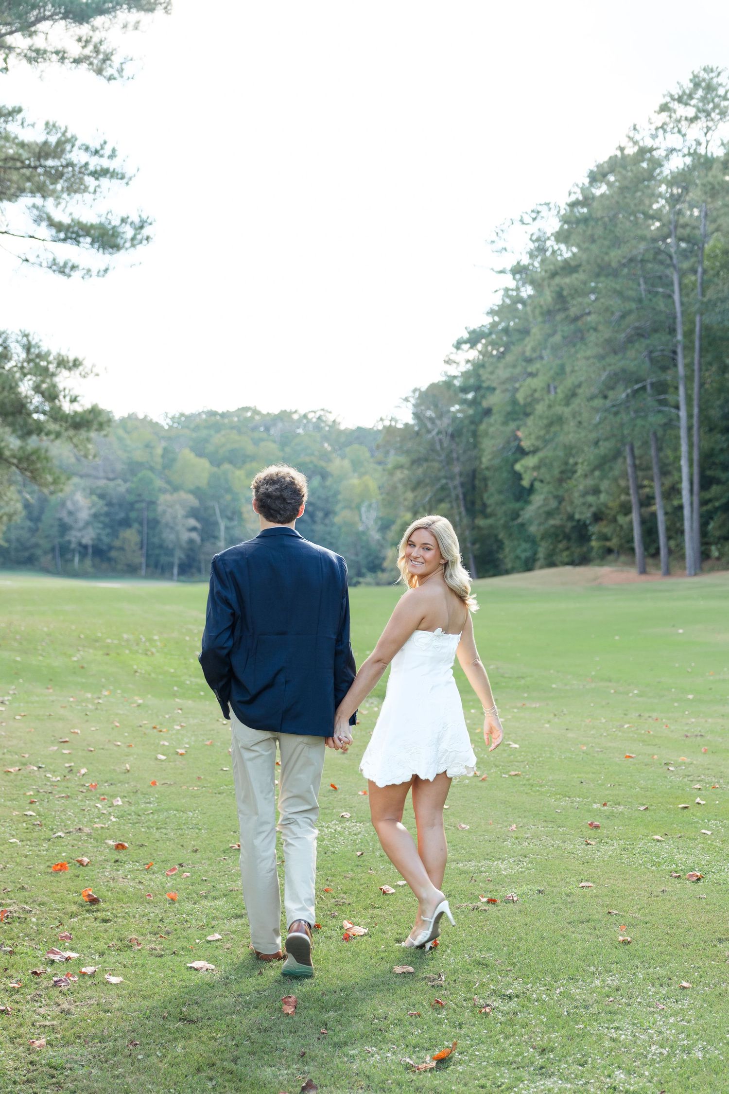 Bride in white dress holds groom's hand walking on green golf course with trees in background.