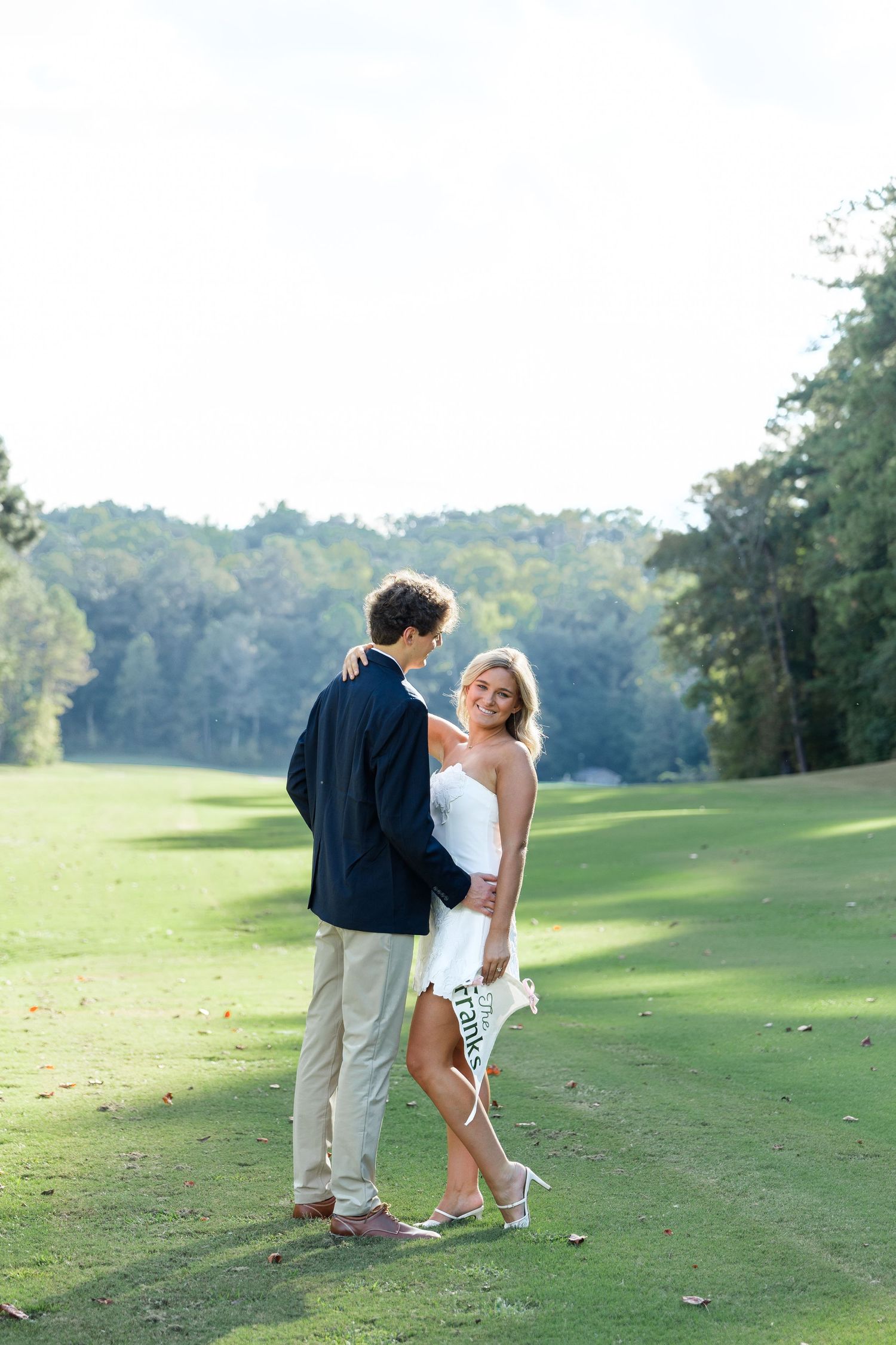 Engaged couple posing on a sunny golf course, bride in white mini dress holding a pennant, groom in navy blazer.