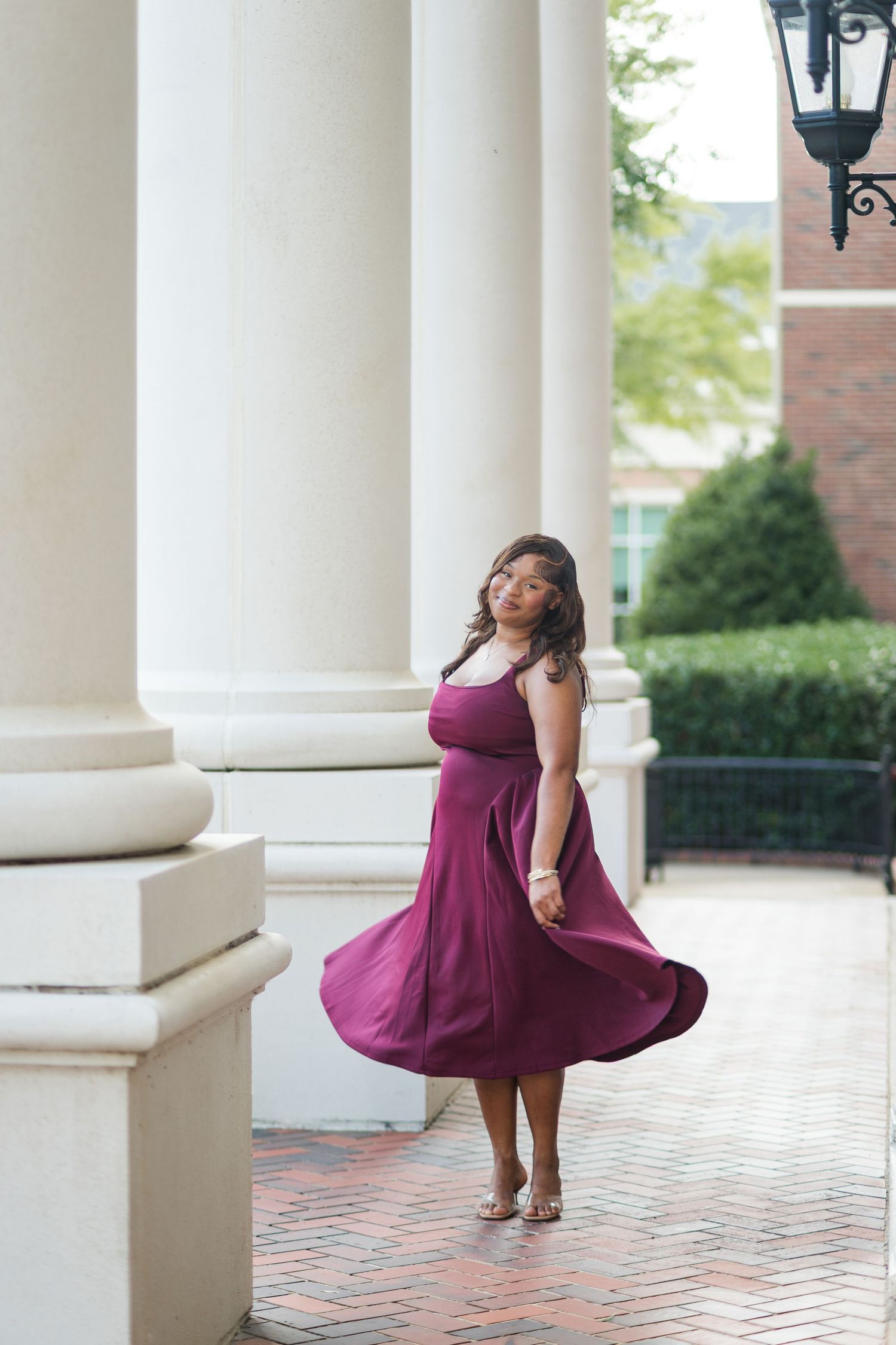 Young woman in a flowing burgundy dress twirling between white columns on a brick pathway.