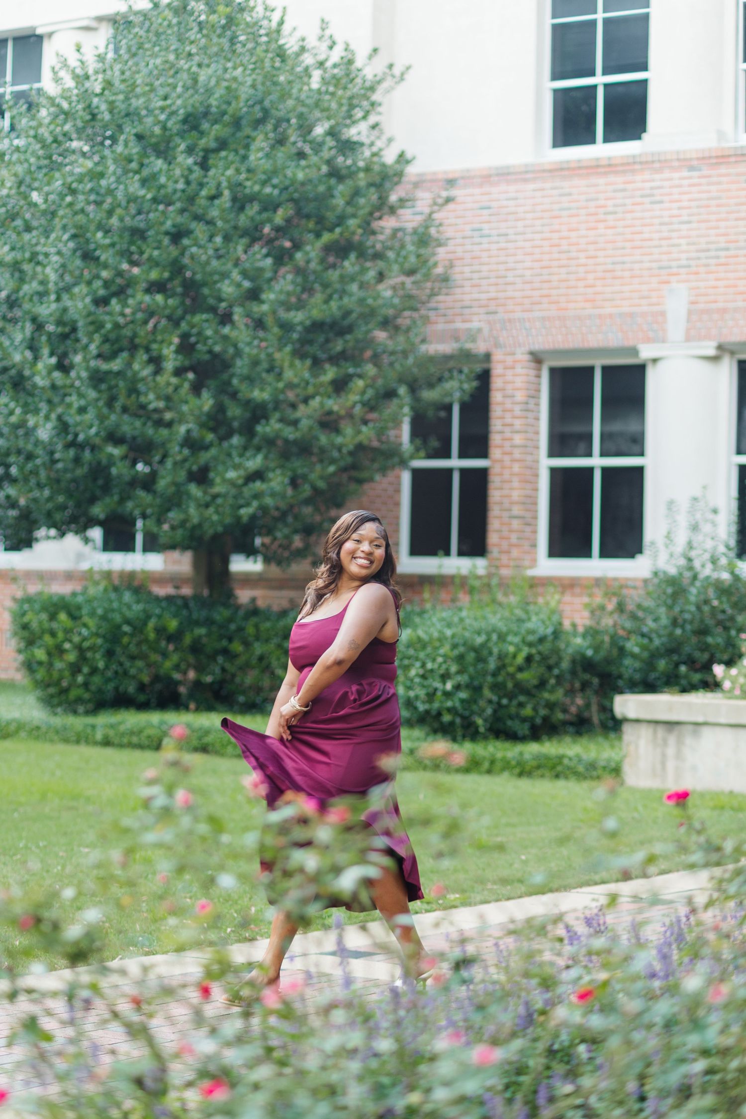 Joyful graduate twirling in burgundy dress among colorful flowers on university campus grounds.