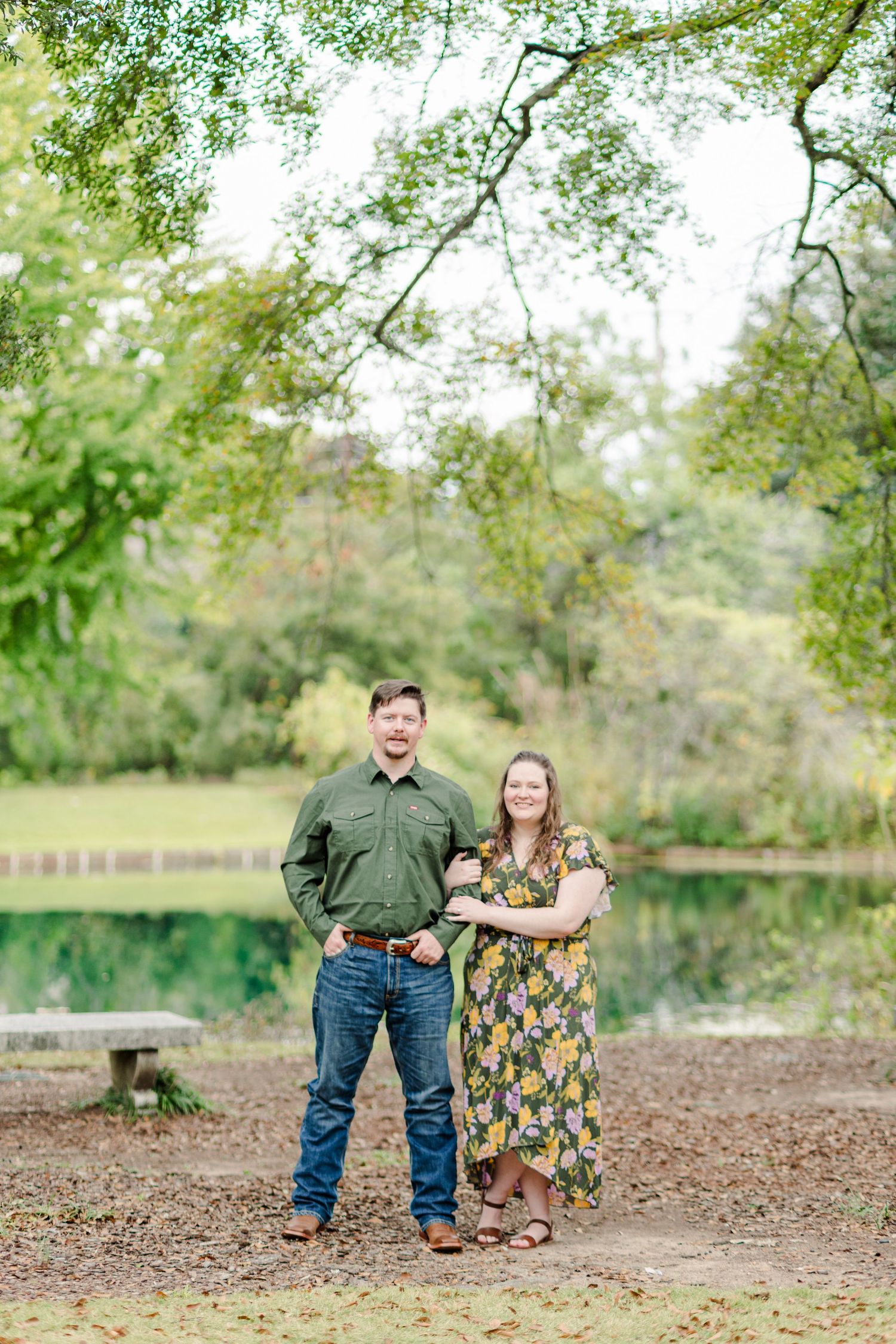 Couple posing together near a serene pond surrounded by lush green trees in an outdoor park setting.