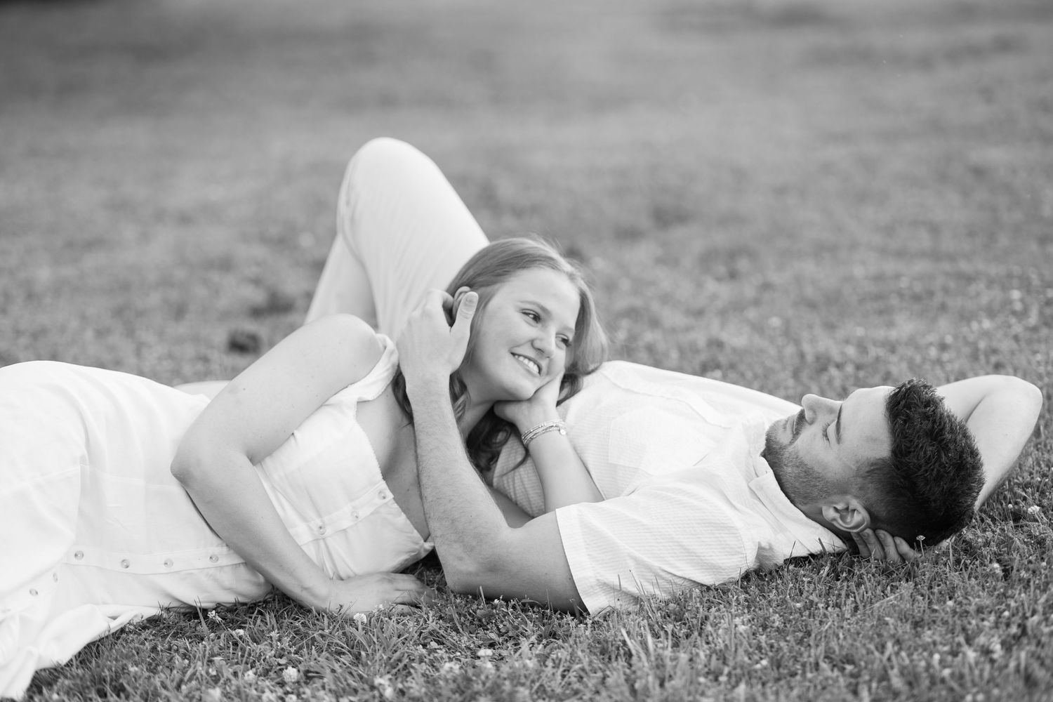 Black and white photo of a couple lying on grass, smiling and gazing at each other romantically.