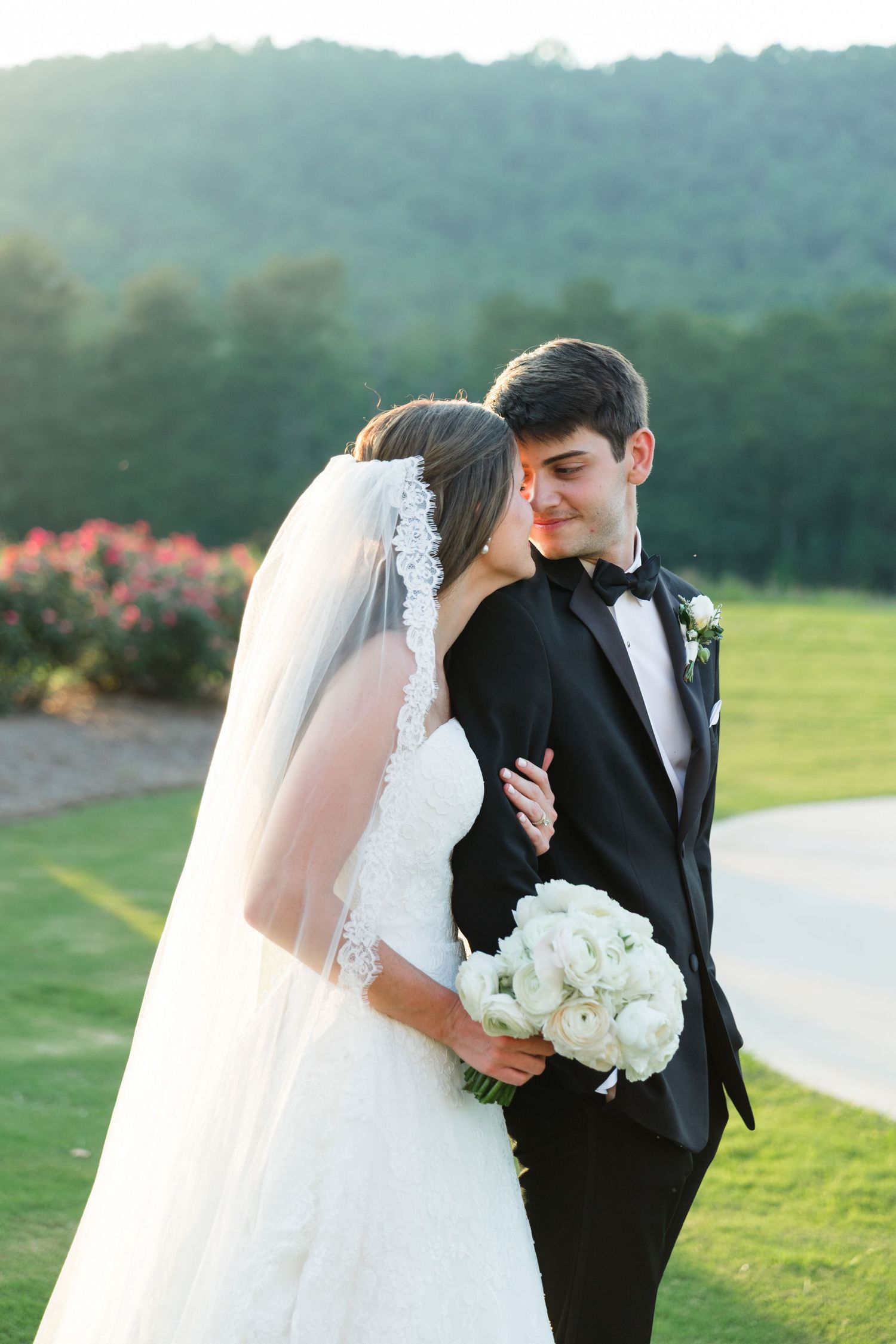 Bride and groom embrace outdoors on green lawn, she holds white bouquet, he wears black tuxedo, lush mountain backdrop.