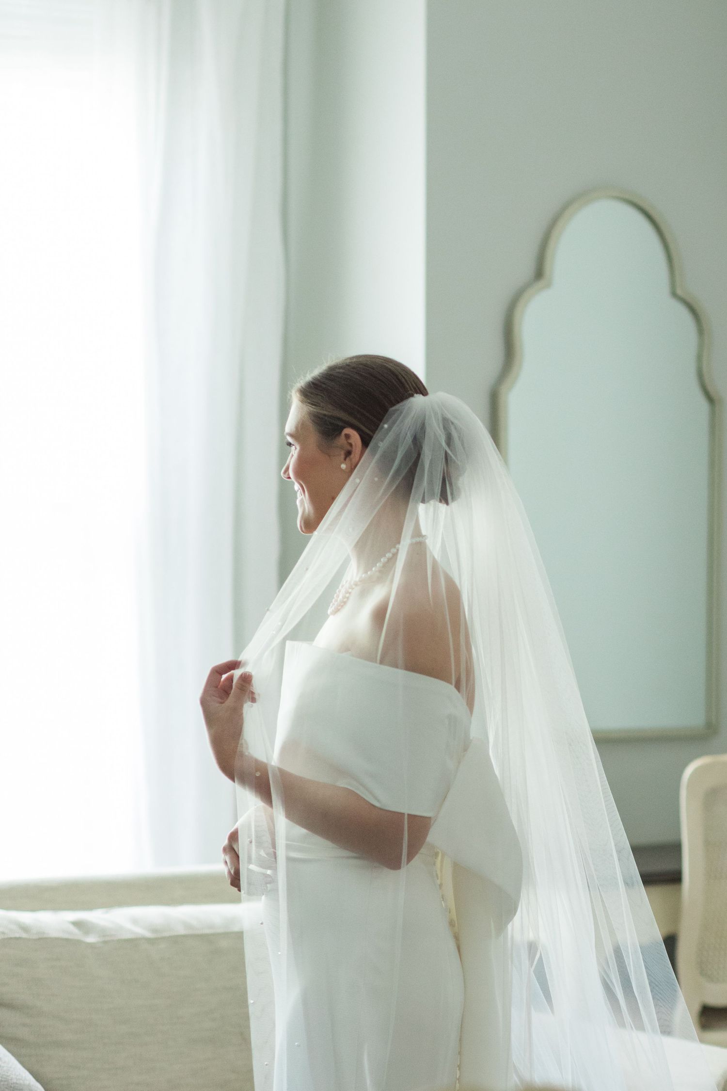 Bride in white off-shoulder gown and flowing veil stands by bright window in elegant room.