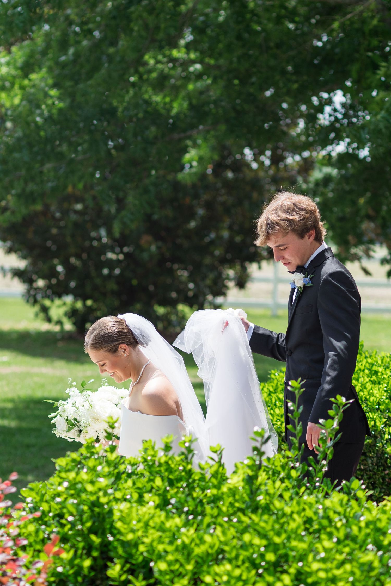 Groom in black tuxedo lifting bride's veil in lush garden during outdoor wedding photos.