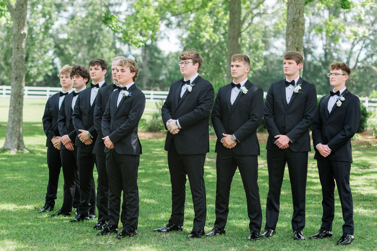 Eight groomsmen in matching black tuxedos with bow ties standing in a row on green lawn.