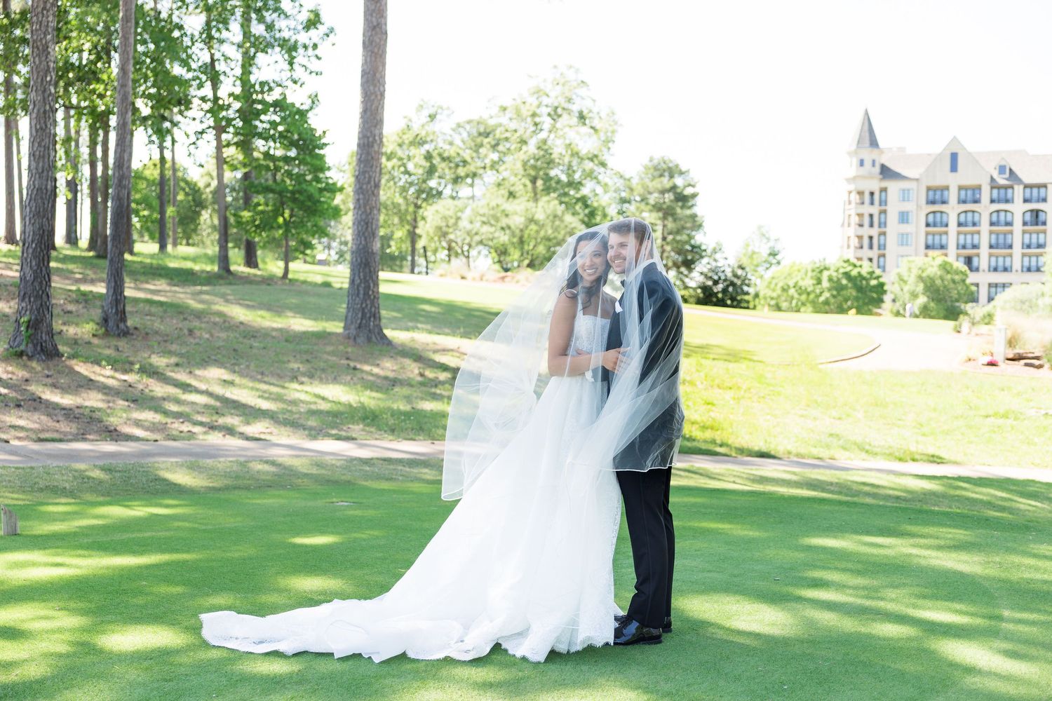 Bride and groom share a romantic kiss on a lush green golf course with a grand estate building in the background.
