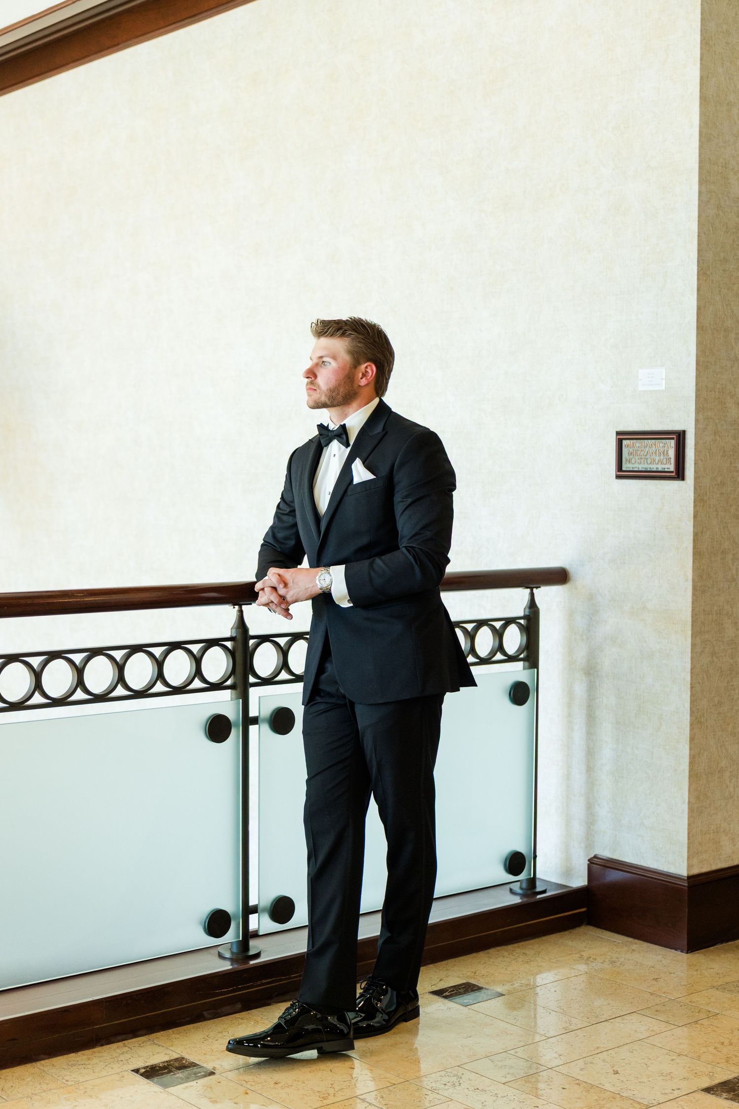Groom in black tuxedo with bow tie stands by glass railing in elegant venue, gazing thoughtfully to the side.