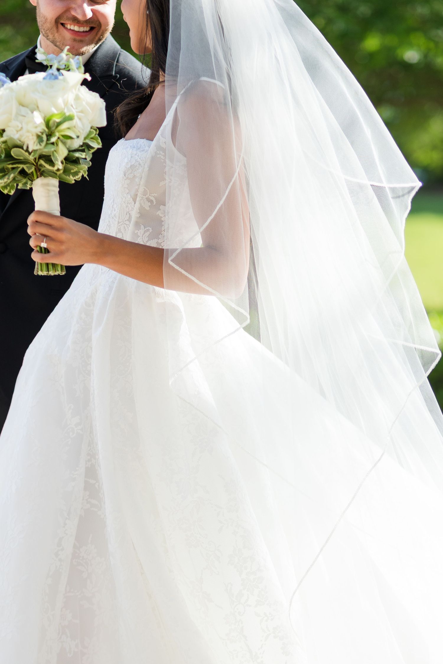 Bride in white lace gown and veil holding bouquet beside smiling groom in black suit outdoors.