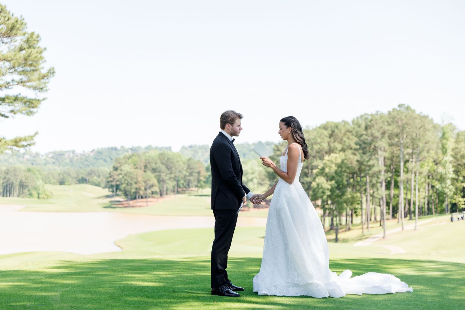 Bride and groom share vows on a lush green golf course surrounded by tall pine trees on a sunny wedding day.