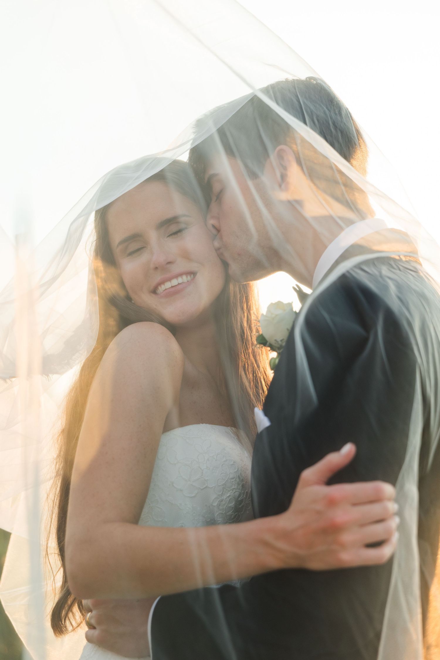 Groom kisses smiling bride under flowing veil in golden backlit sunset portrait.