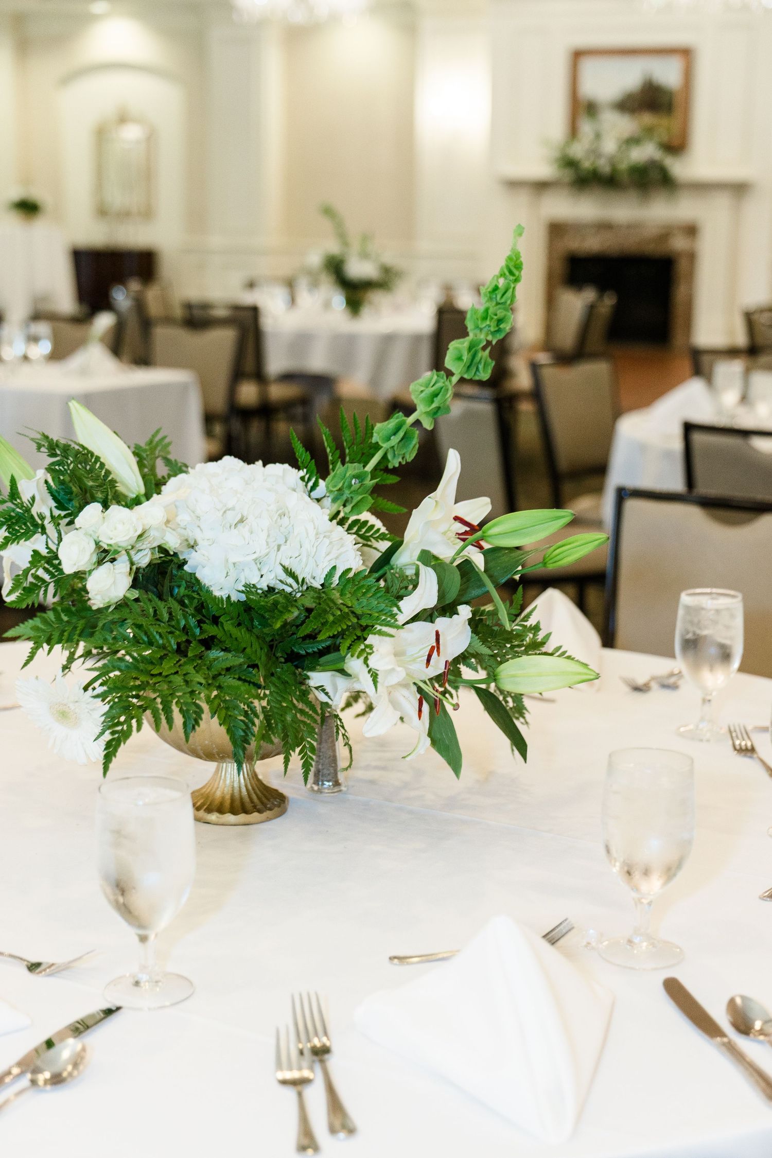 Elegant wedding reception table with white hydrangea and green fern floral centerpiece in a gold vase.