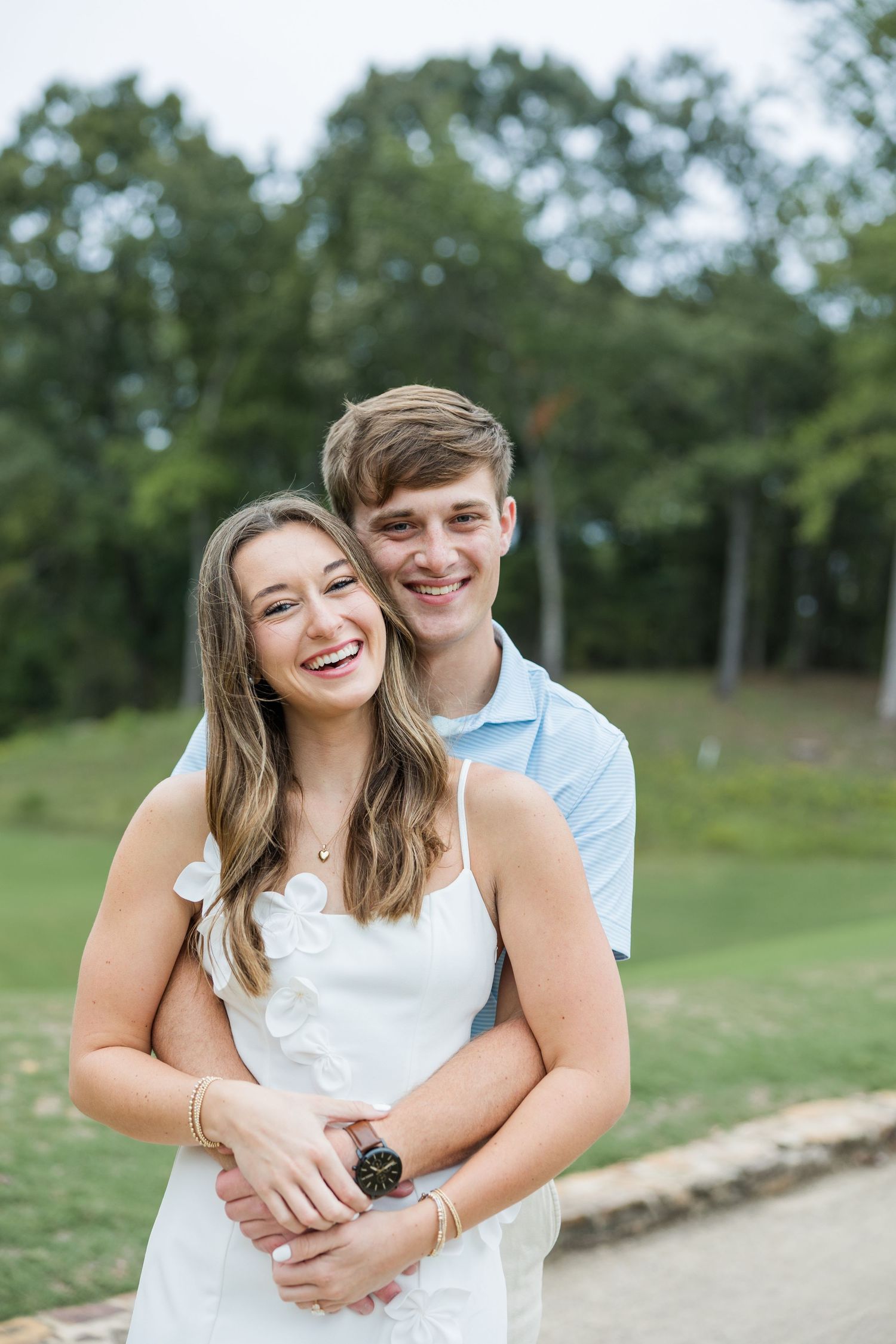 Smiling woman in a white dress leans back into her fiancé's embrace during an engagement photo session with trees in the background.