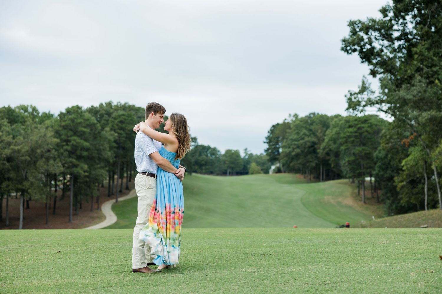 A couple in a loving embrace stands on a grassy golf course fairway surrounded by green trees under an overcast sky.