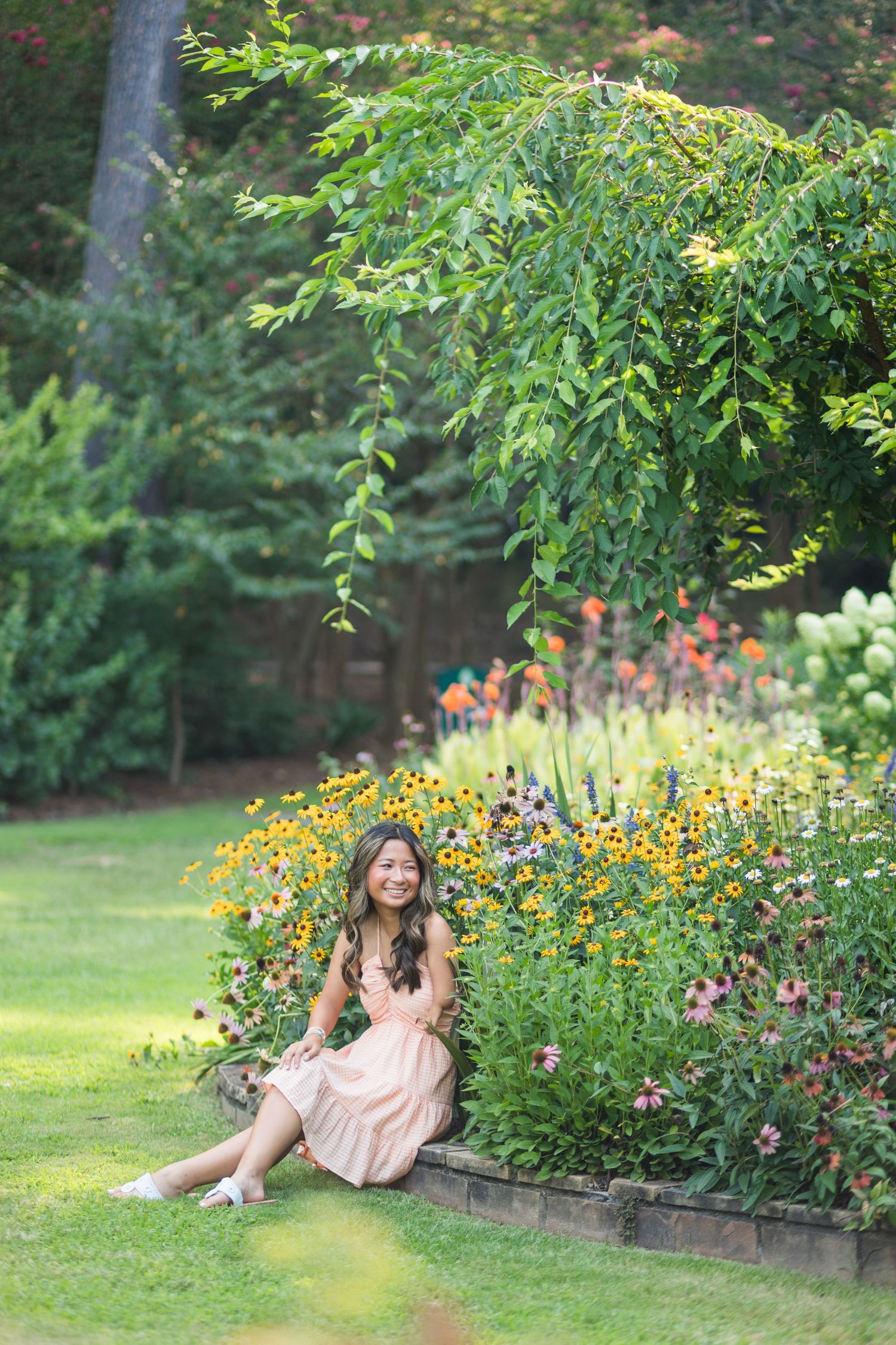 A young woman in a peach dress sits relaxed beside blooming black-eyed Susans in a lush garden.