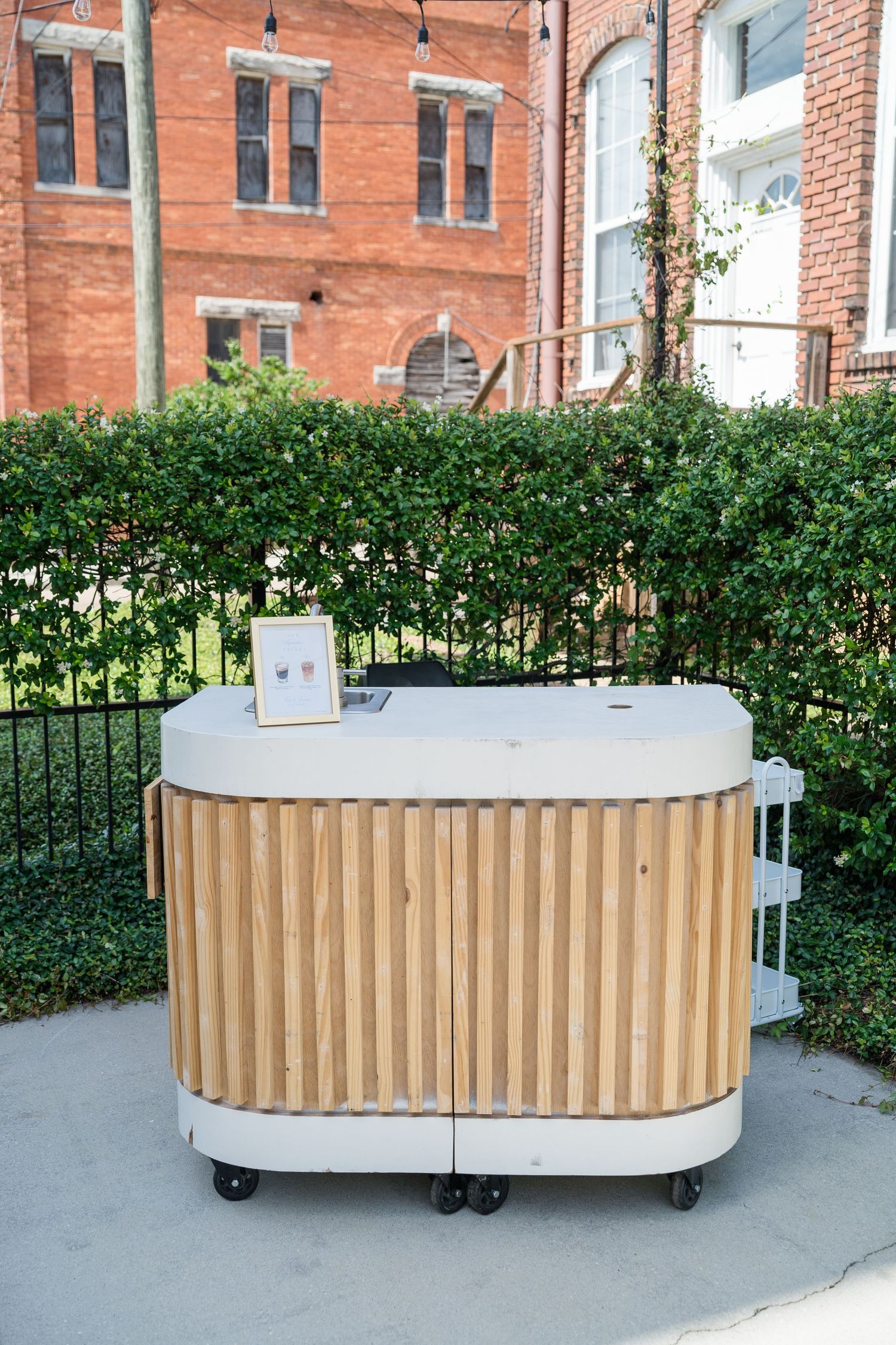 Elegant portable wooden bar cart with white top and casters, set outdoors against lush green hedges at a wedding venue.