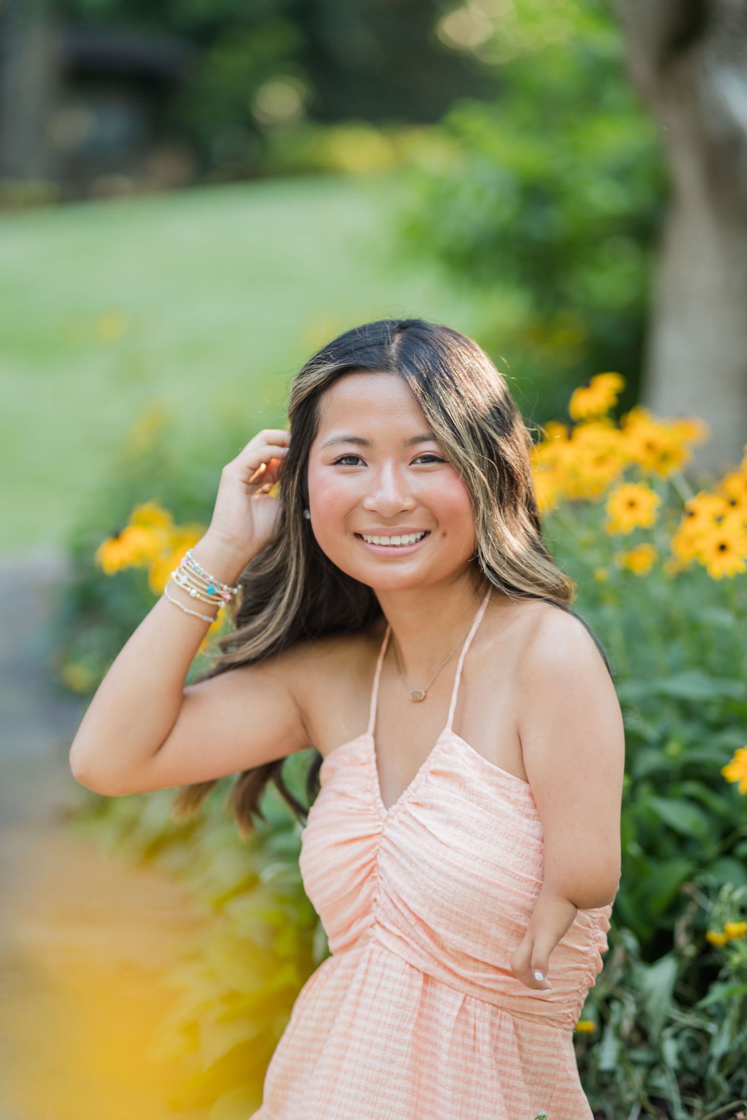 Young woman in peach sundress smiling outdoors among yellow flowers in a lush green garden.