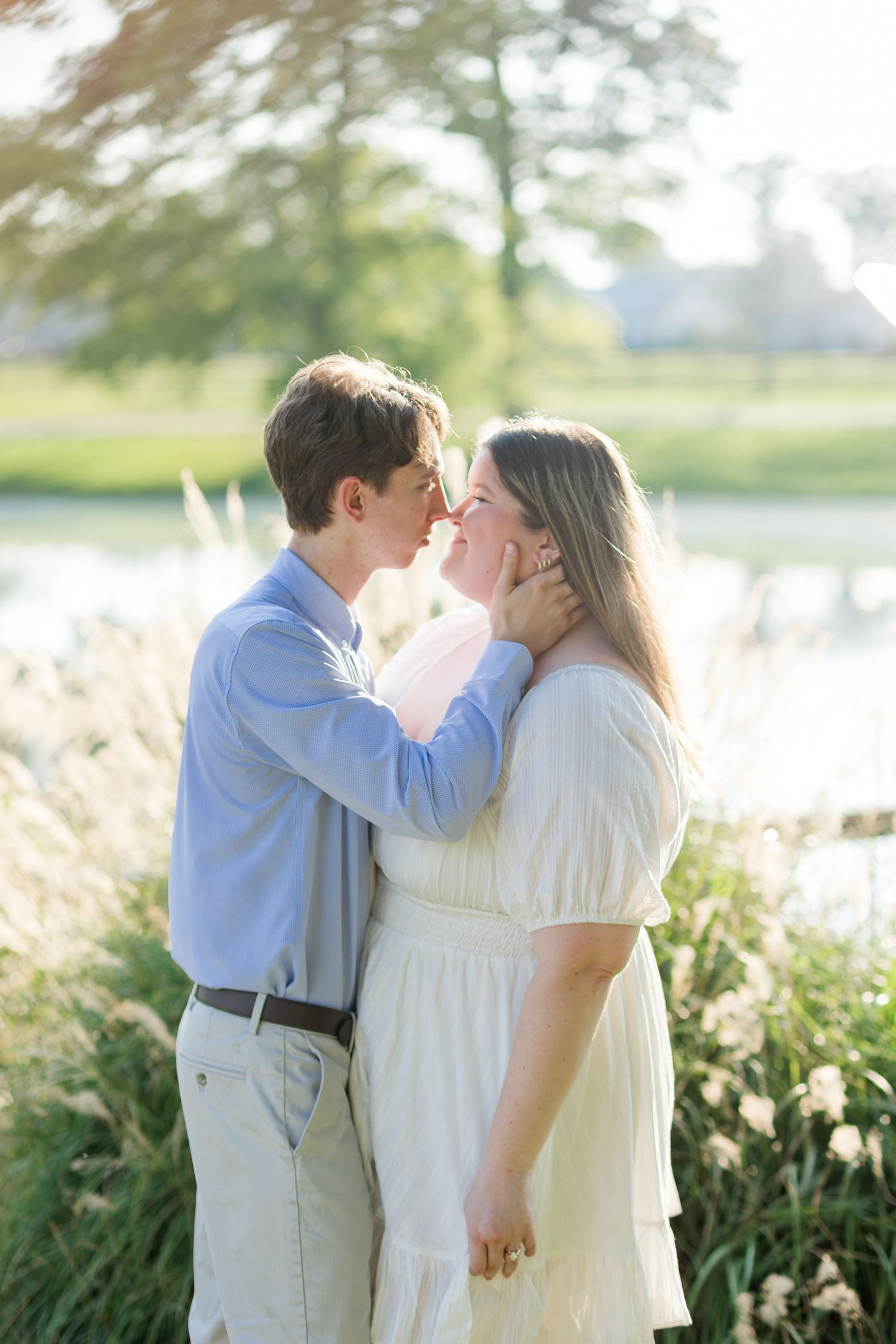 A couple shares a tender moment near a pond, the man in blue cradling her face as they lean in for a kiss.