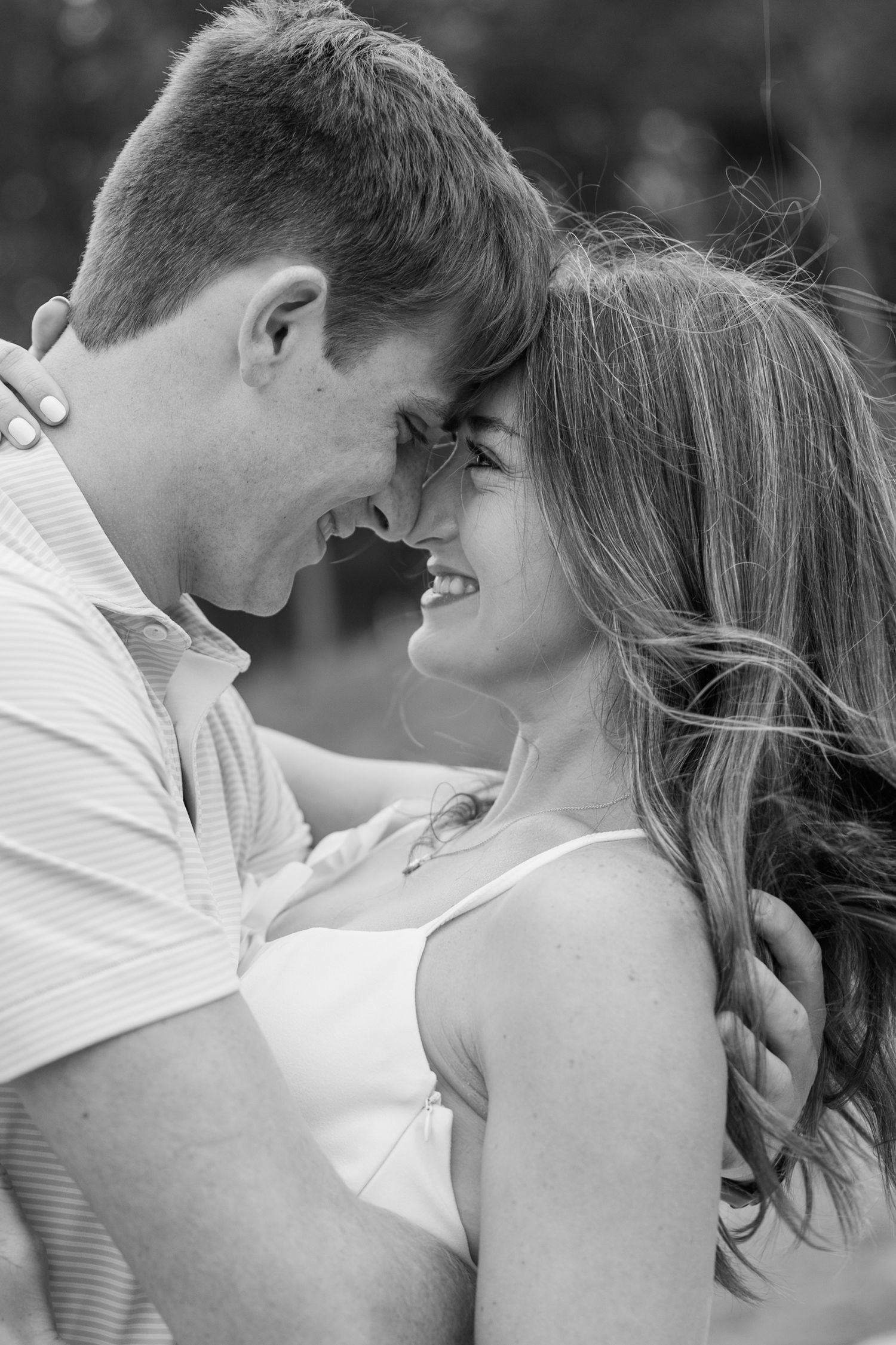 Black and white engagement photo of a happy couple embracing with foreheads touching and smiling, her hair blowing in the breeze.