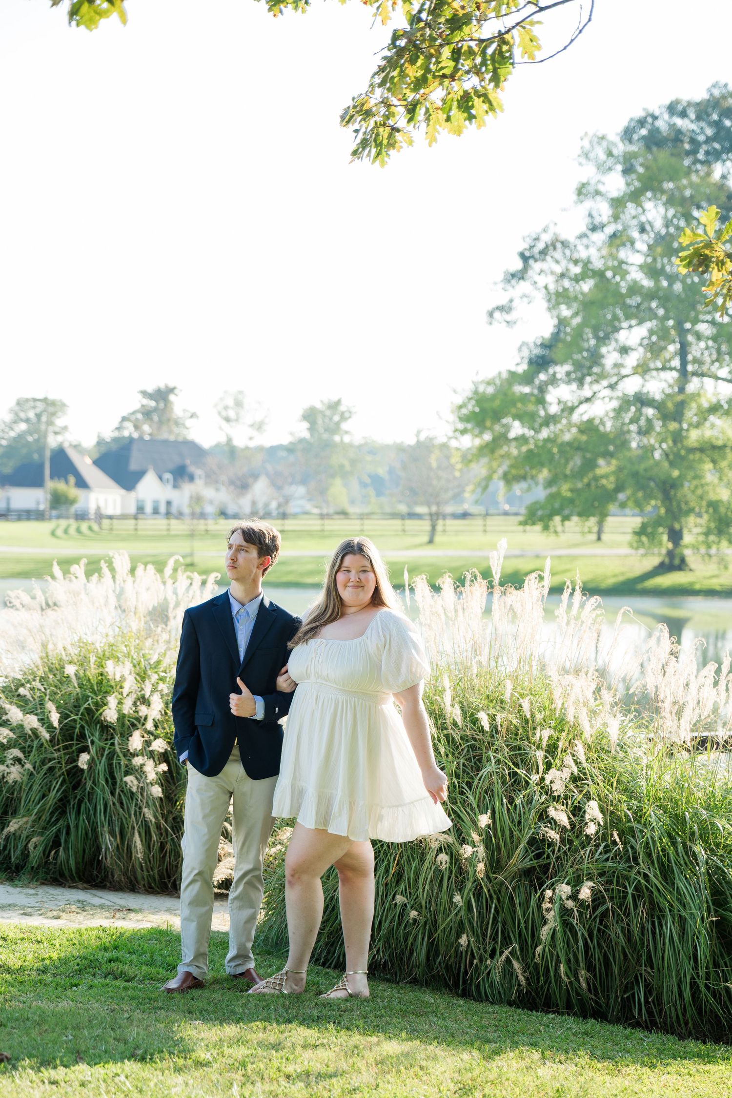 Couple posing outdoors near a pond with tall ornamental grasses, man in navy blazer, woman in white dress.