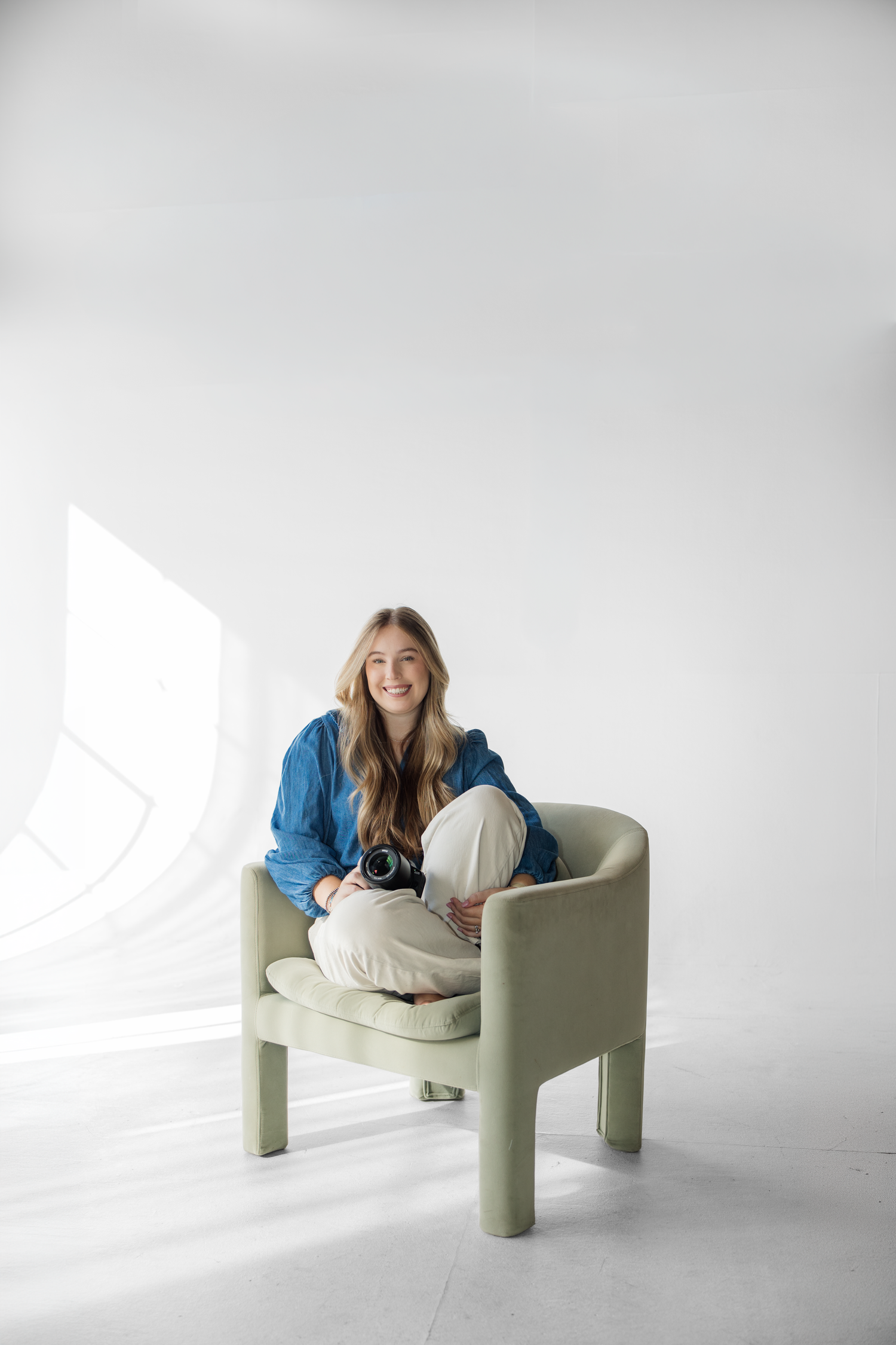 Smiling woman in blue shirt sitting cross-legged on a modern sage green armchair in a bright white studio.