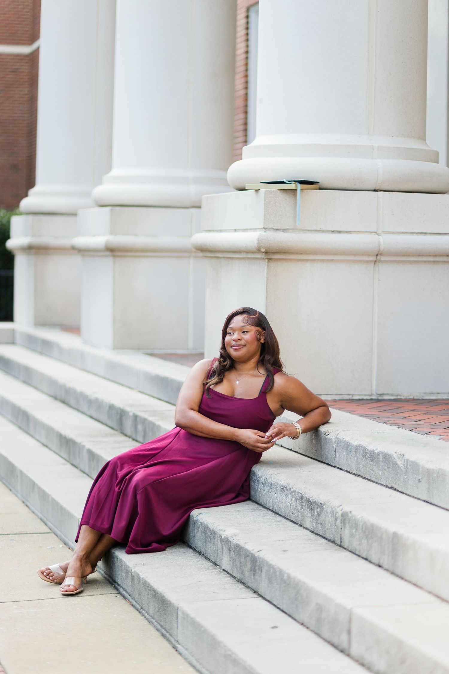 Graduate in burgundy jumpsuit relaxing on steps beside white columns of university building.