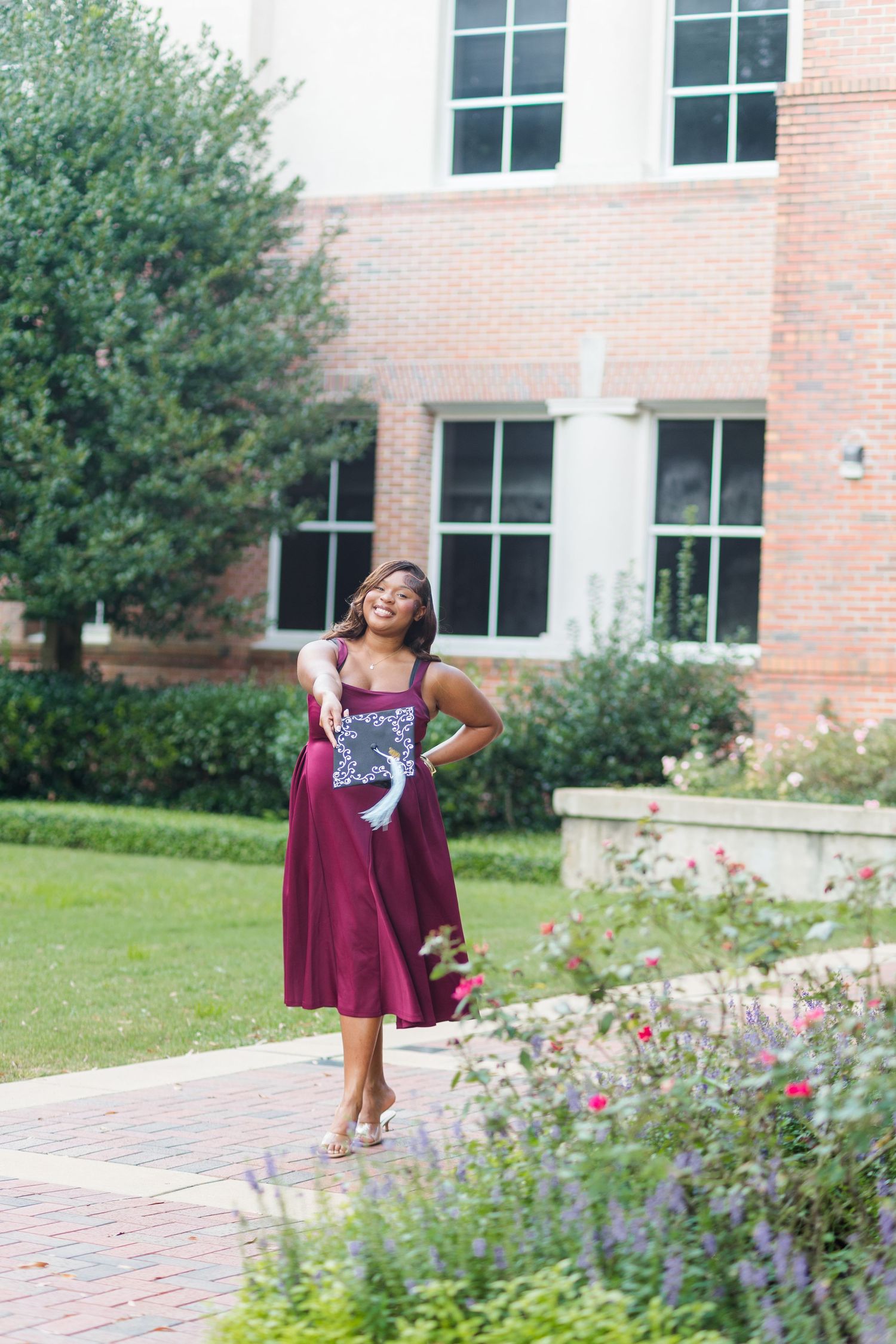 Graduating student in burgundy dress holding decorated cap while walking through campus garden path.