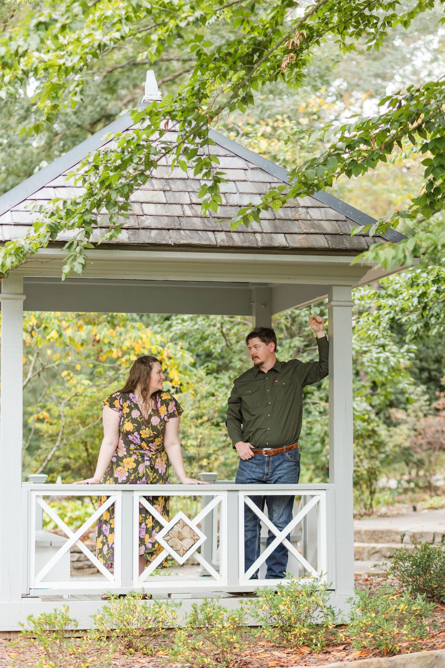 Couple smiling at each other in a white gazebo surrounded by lush green trees during an outdoor engagement session.