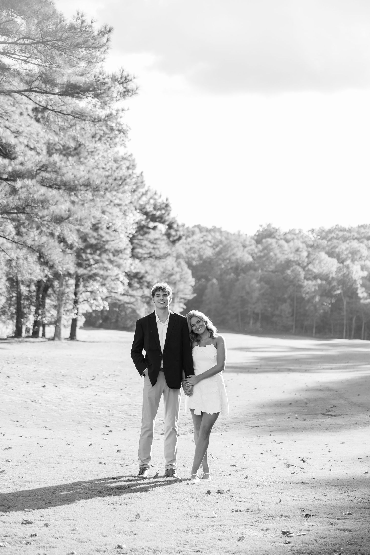 Black and white photo of a couple holding hands and walking along a sandy path surrounded by tall pine trees.