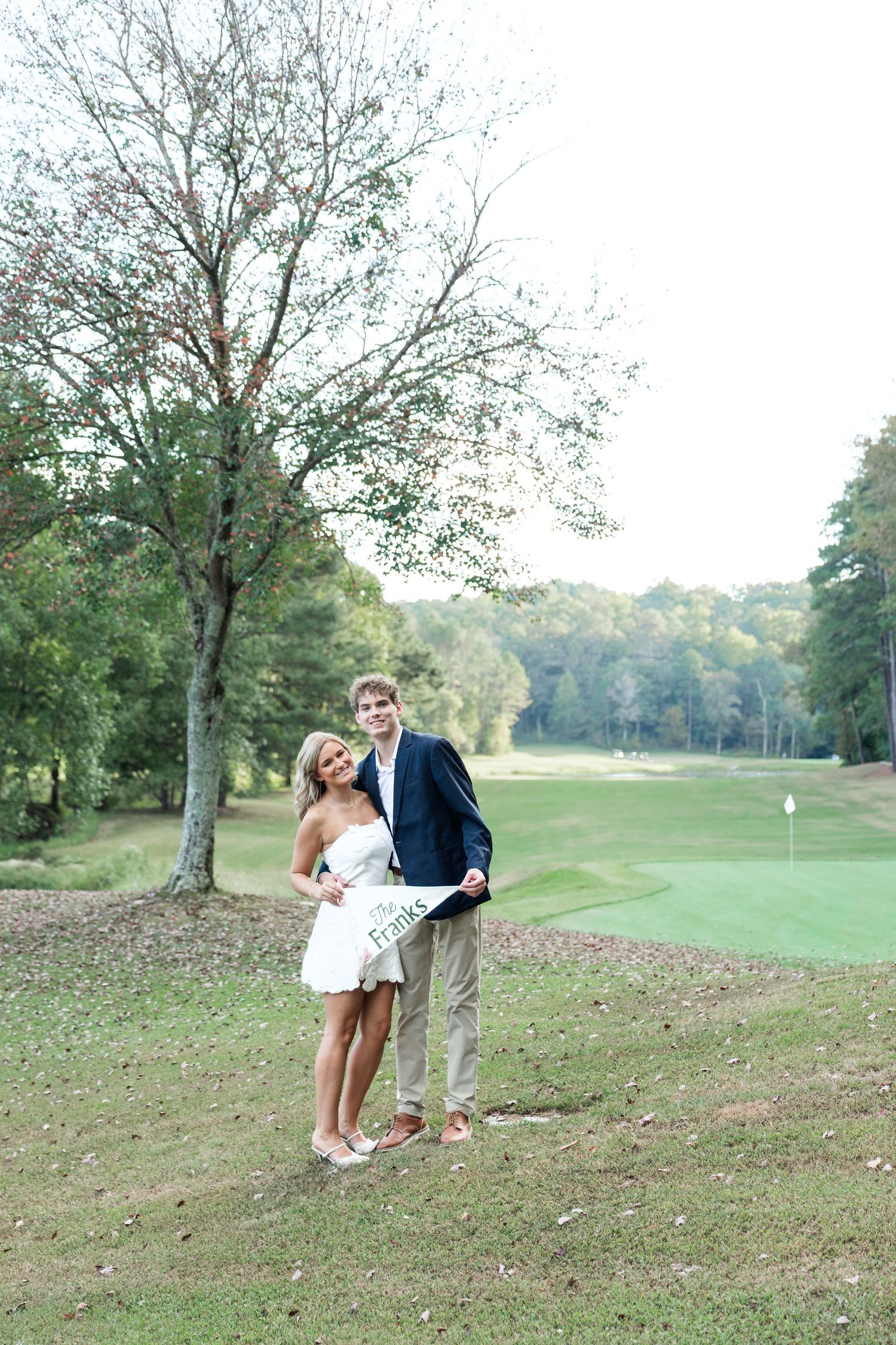 Newlyweds posing on golf course holding The Franks pennant sign, woman in white dress and man in blazer.