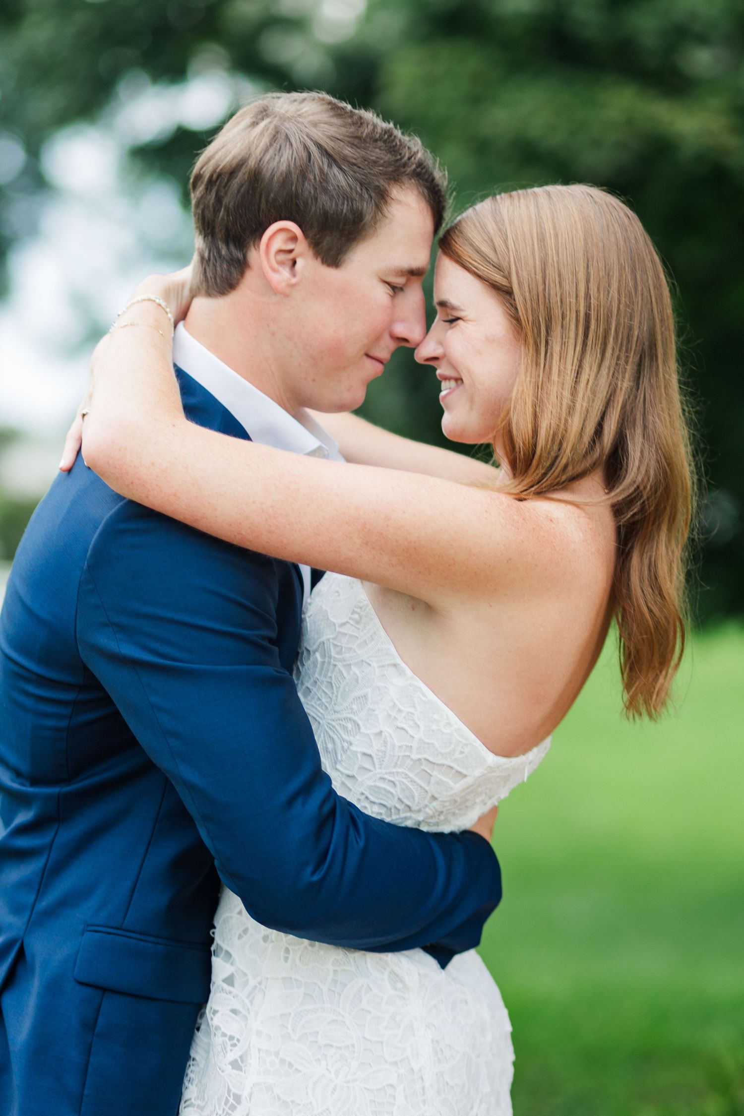 Bride and groom sharing an intimate forehead touch moment, smiling, outdoors with lush green background.
