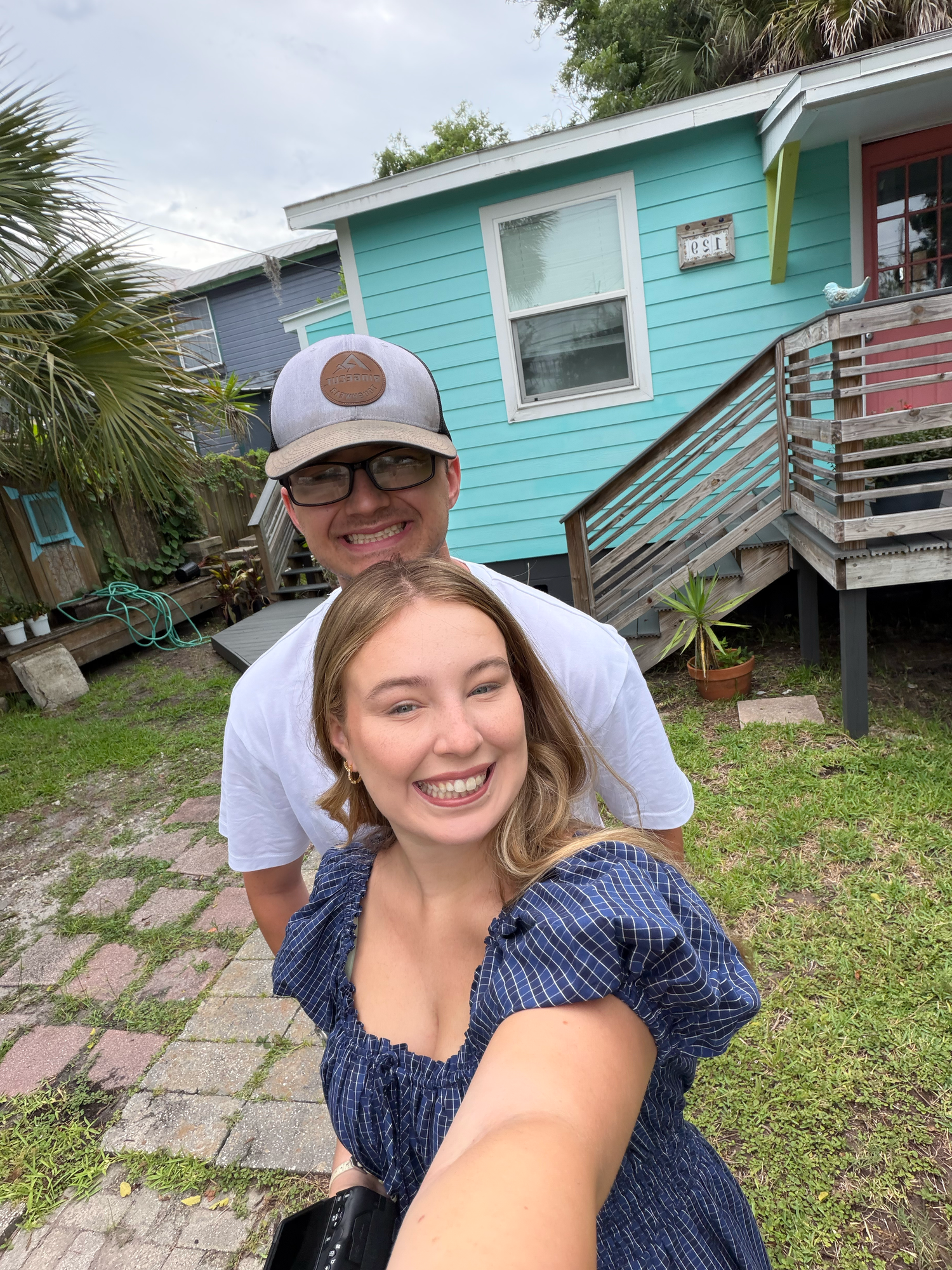Smiling couple taking selfie in front of colorful turquoise beach house on sunny day.