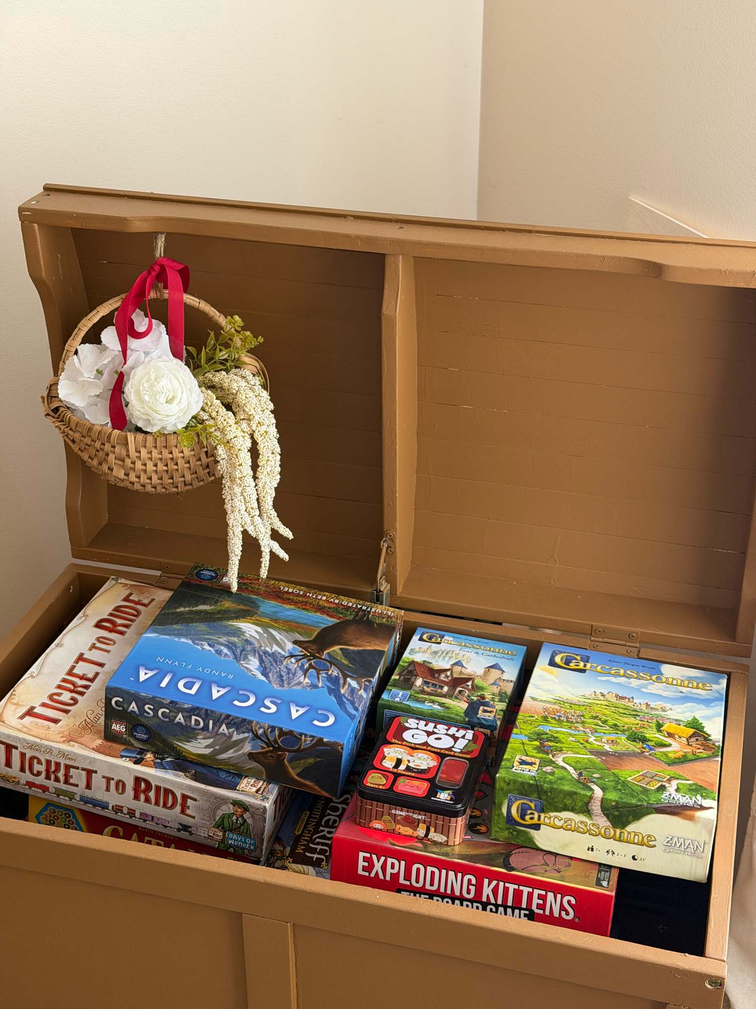 Wooden storage chest filled with popular board games including Ticket to Ride, Cascadia, and Exploding Kittens.