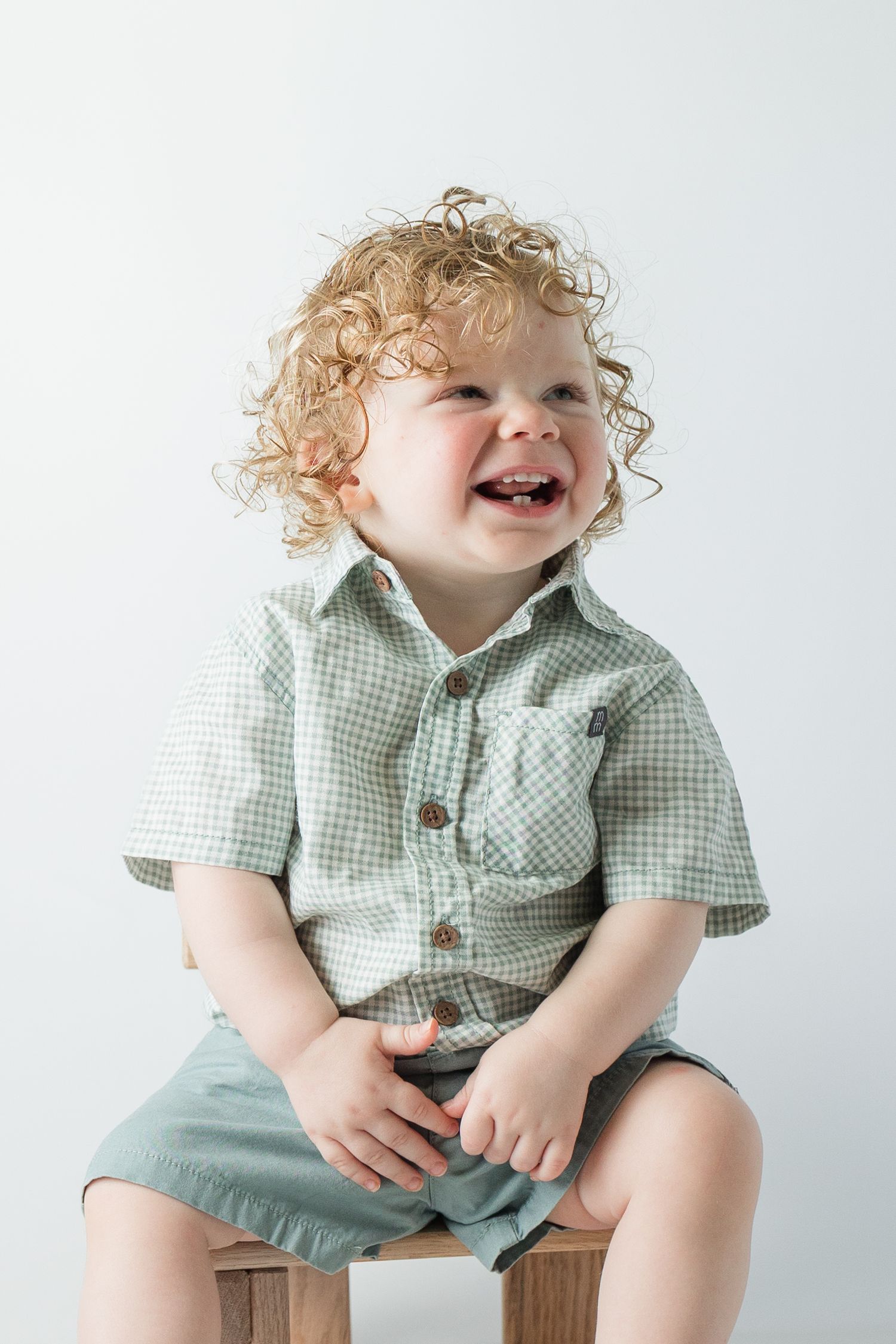 Happy curly-haired toddler boy laughing while sitting on a wooden stool against white background.
